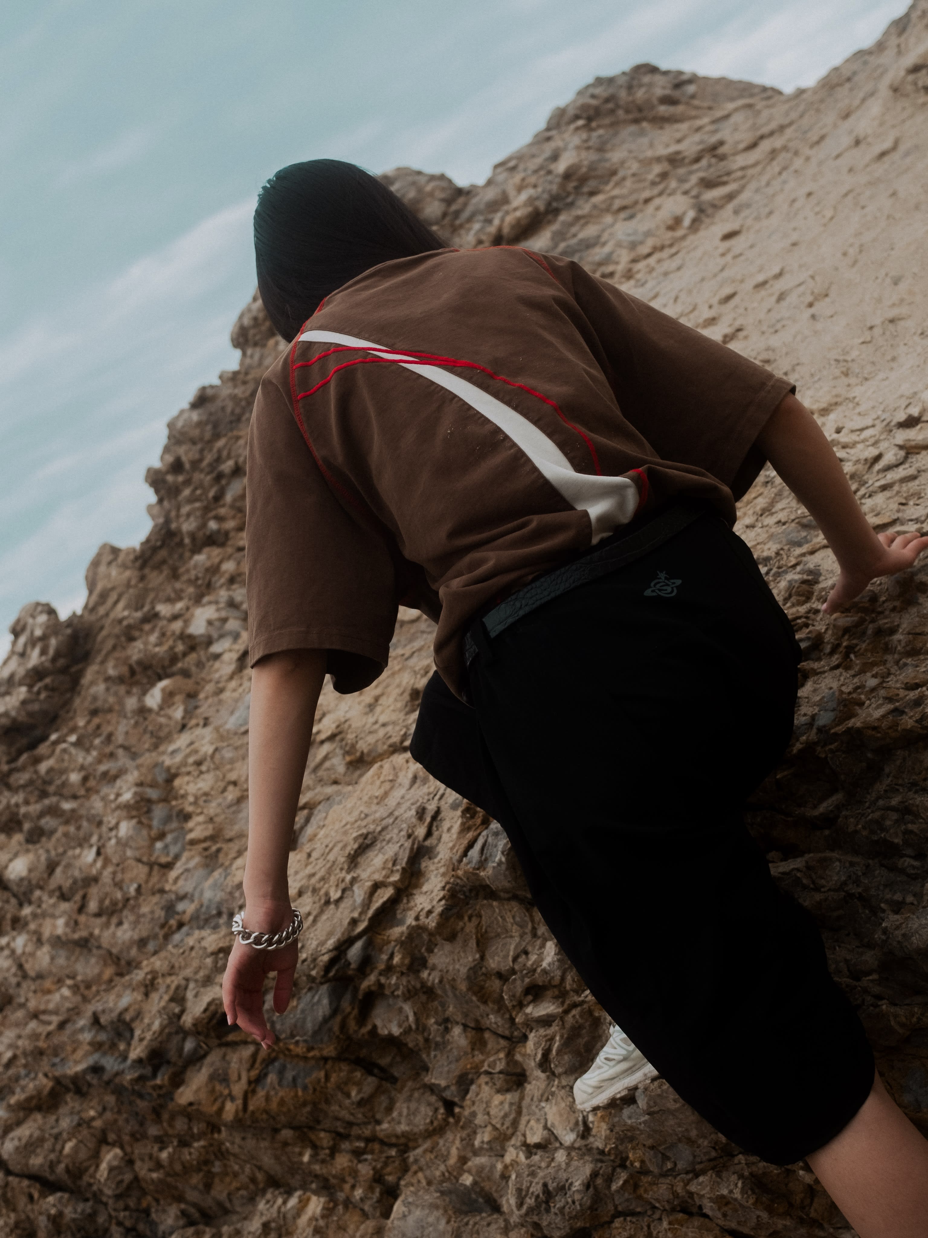 A person in a brown t-shirt and black pants climbs a rugged, rocky cliffside under a pale blue sky.