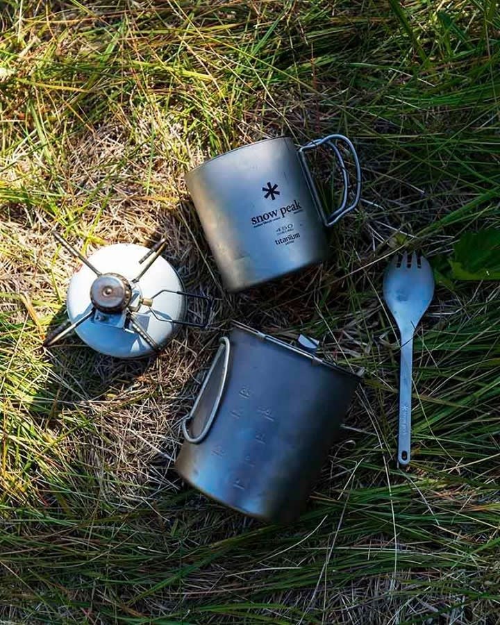 Camping cookware, including a stove, mug, pot, and spork, rests on a bed of green and yellow grass.