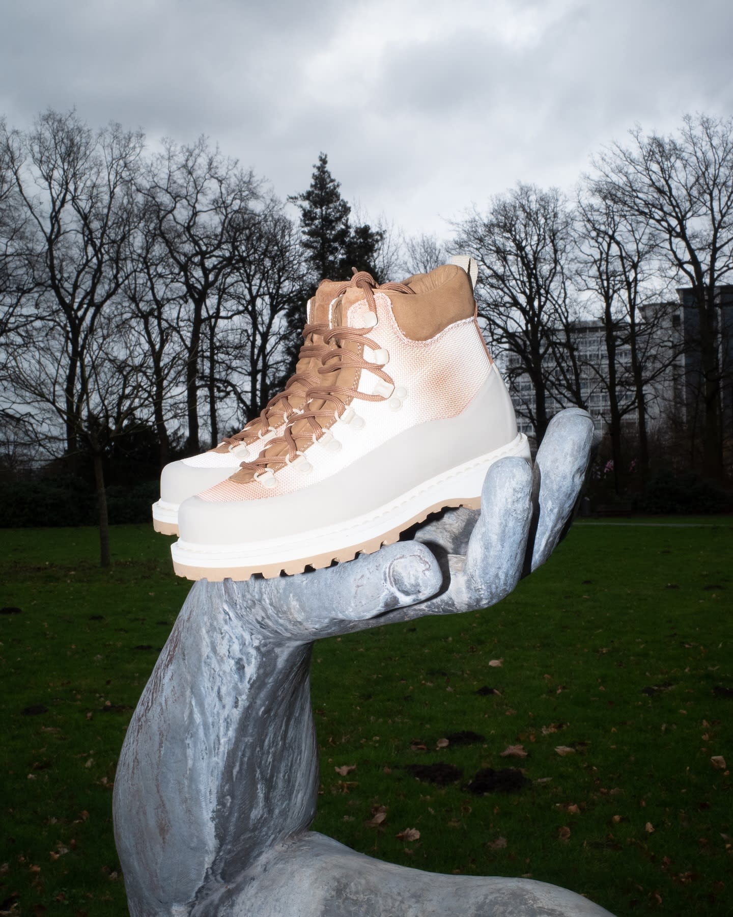A pair of light-colored hiking boots rests on a large, silver, open hand sculpture in the foreground, with a park landscape featuring bare trees and buildings in the background.