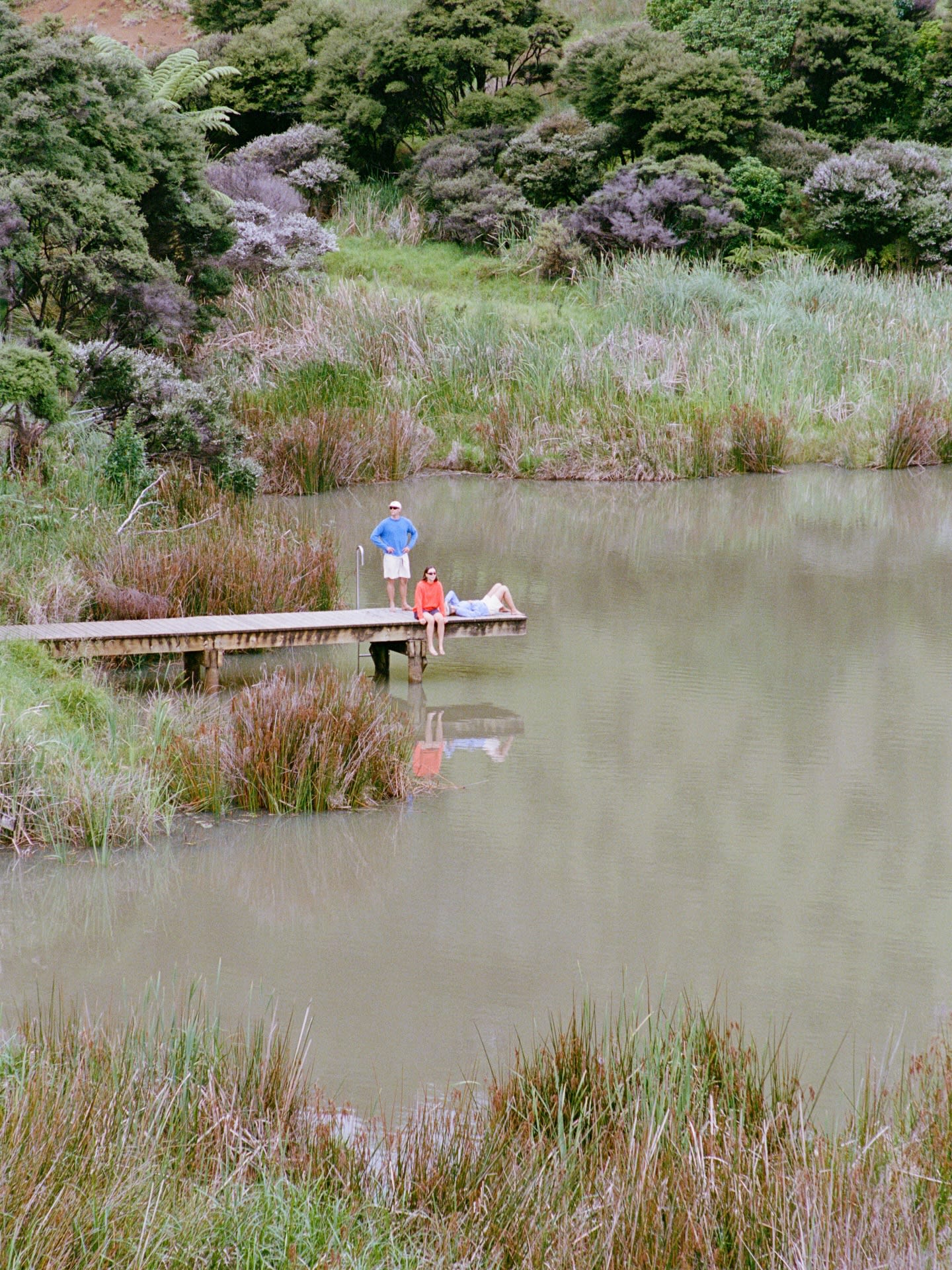 Two people relax on a wooden pier extending into a calm pond, surrounded by lush green grass and dense forest.