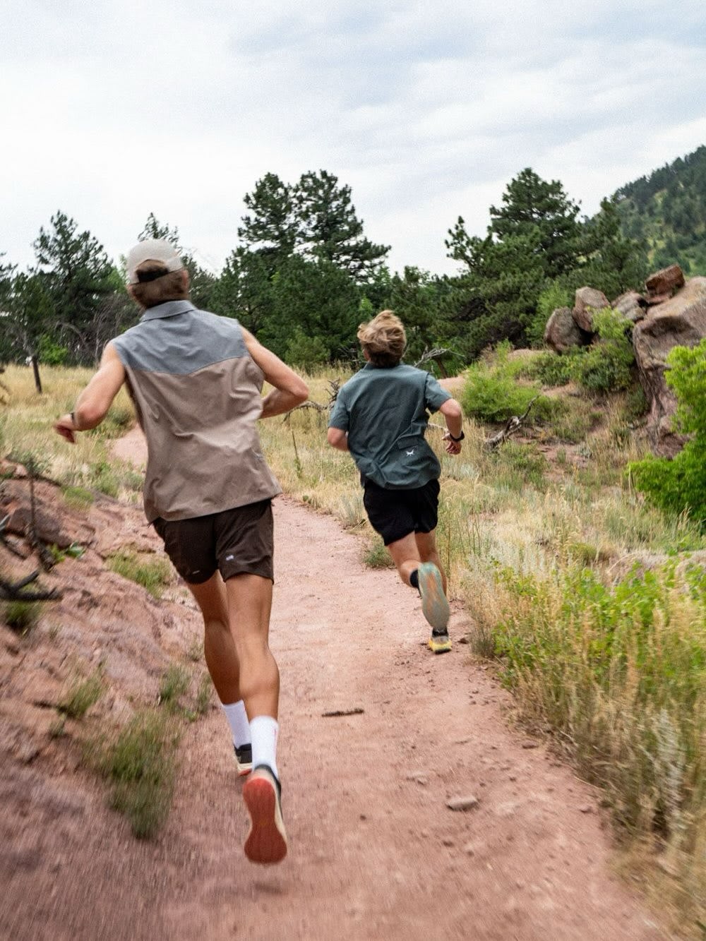 Two men run along a dirt trail through a hilly landscape filled with pine trees and rocky outcrops.