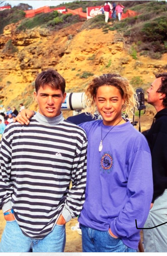 Two young men stand side-by-side in the foreground against a backdrop of a sandy cliff and a grassy hill.