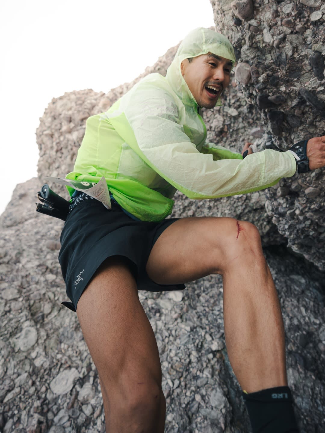 A man in athletic wear climbs a rocky incline, with a bright sky visible in the background.