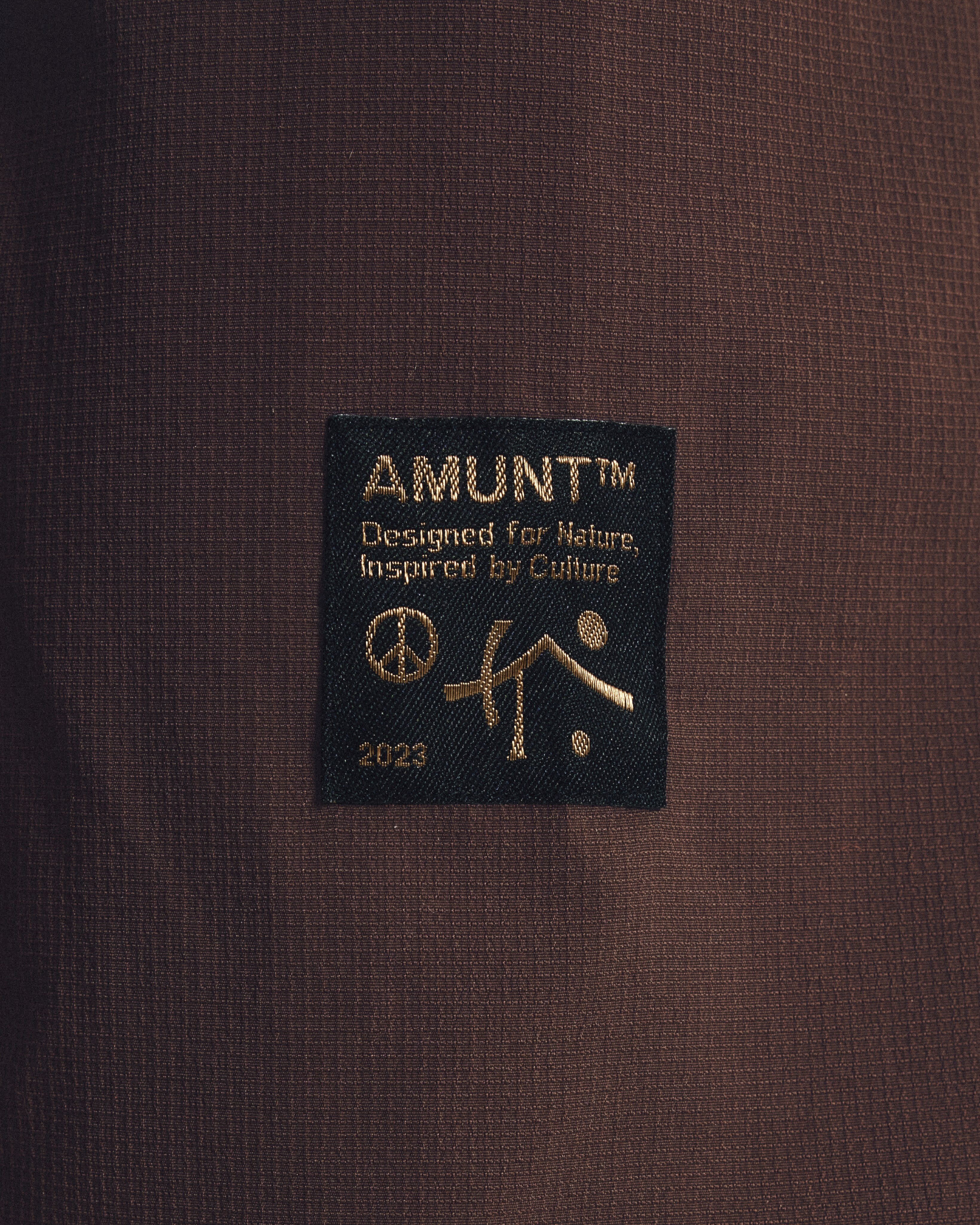 A black fabric label with gold text and symbols is sewn onto a dark brown textured garment.