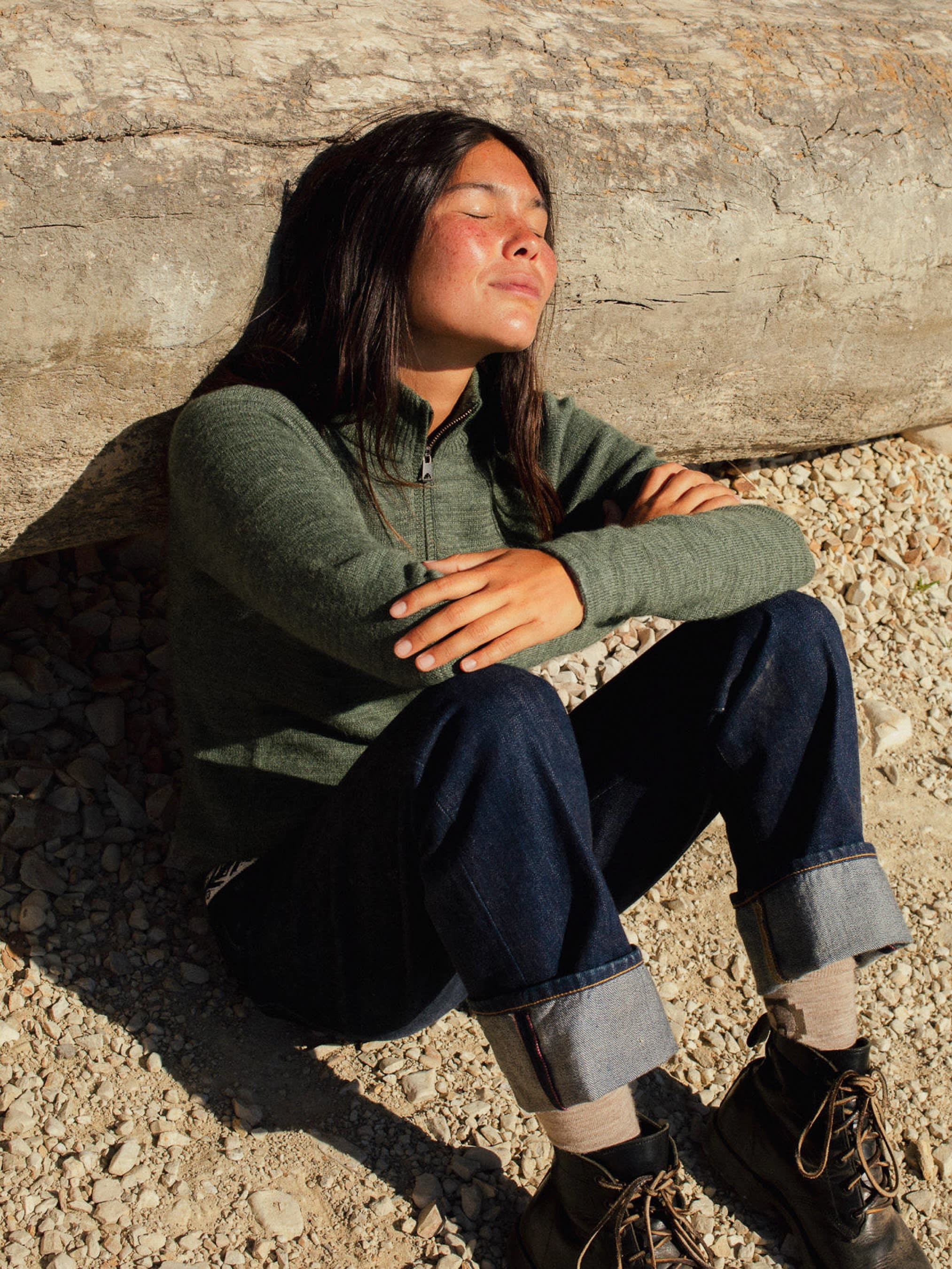 A young woman with her eyes closed and arms crossed sits on a rocky ground in front of a large, weathered log.