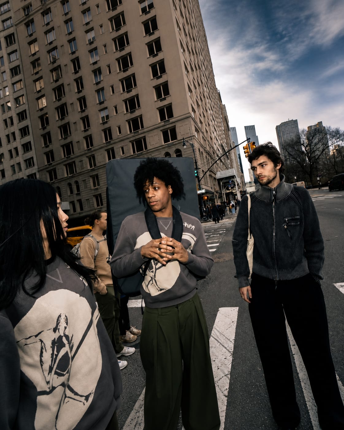 Three young people stand on a crosswalk in front of a tall, modern building under a cloudy sky.