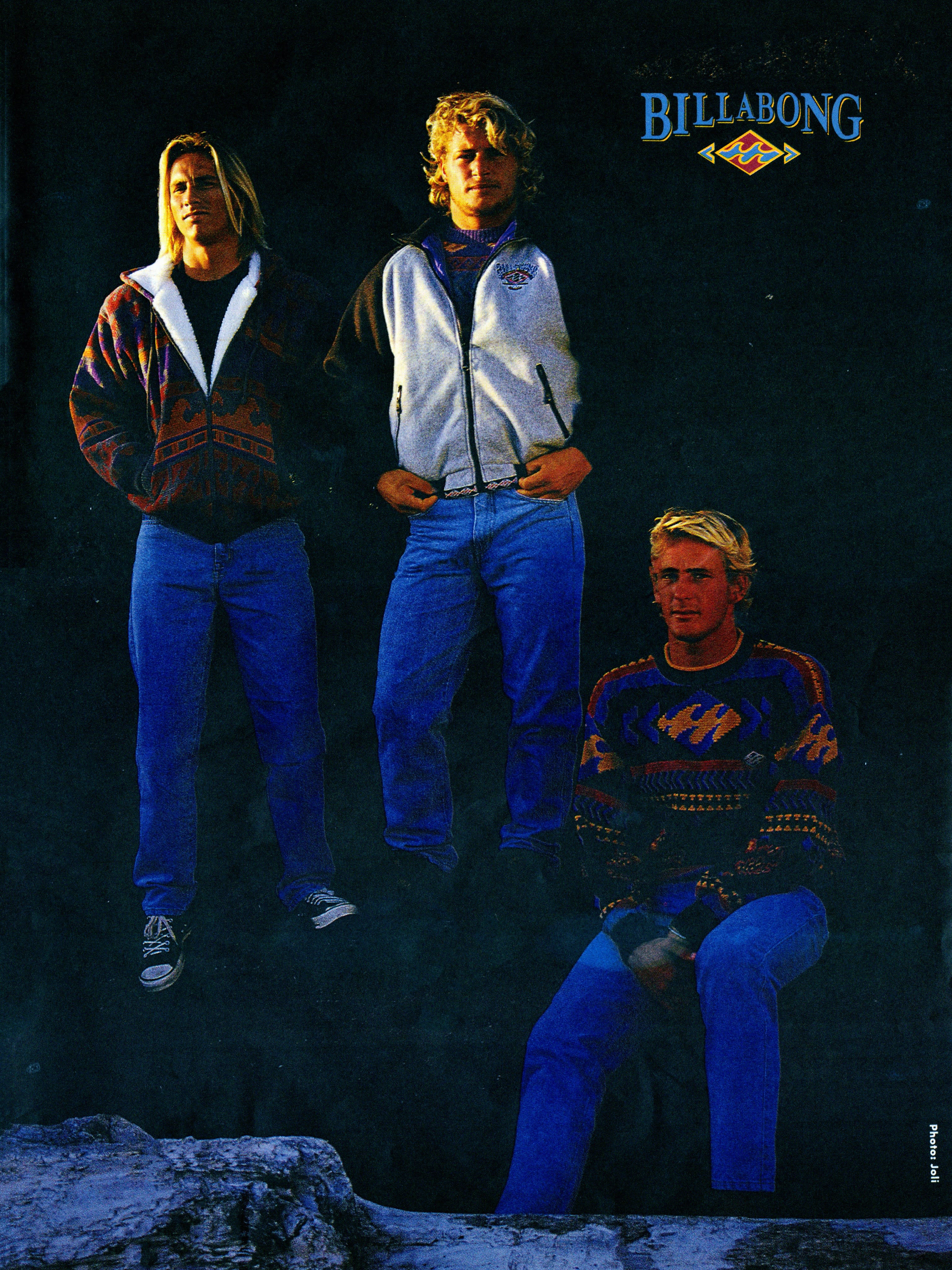 Three men wearing Billabong apparel and blue jeans pose against a dark, textured background with rocky terrain in the lower foreground.
