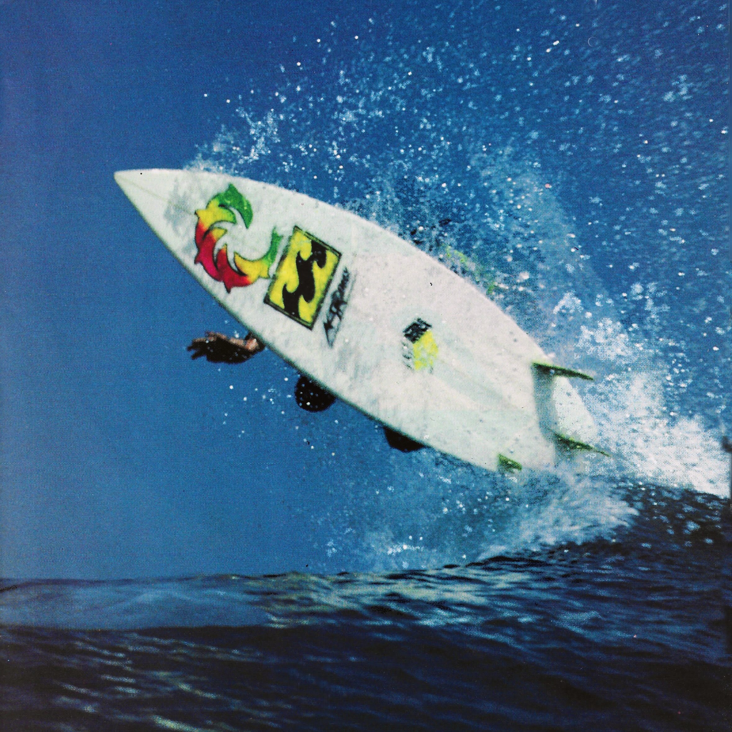 A surfer catches air on a white surfboard amidst a spray of water against a clear blue sky and ocean.