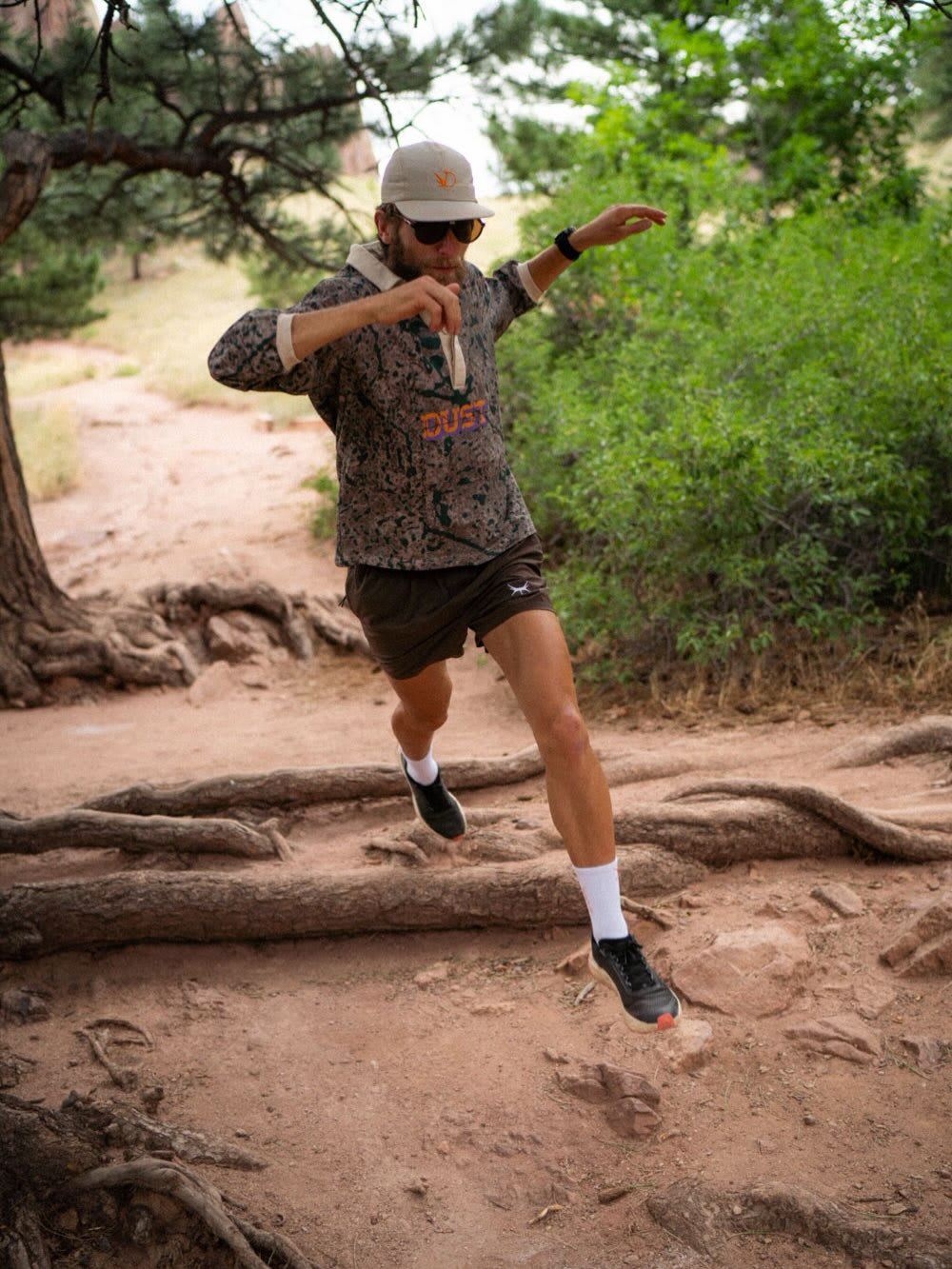 A man in a patterned shirt and cap balances on one leg atop a sandy path surrounded by tree roots and green foliage.