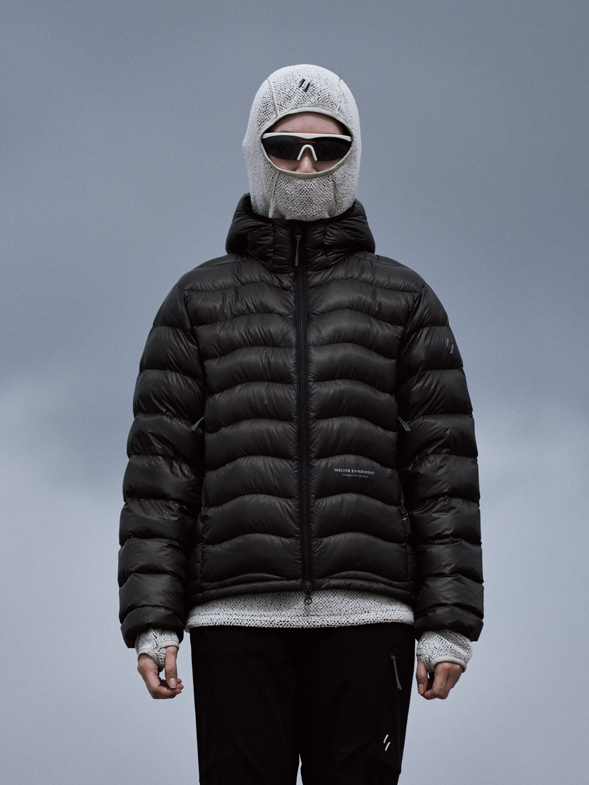 A person wearing a black puffer jacket, black pants, a white knit balaclava, and sunglasses stands against a solid grey background.