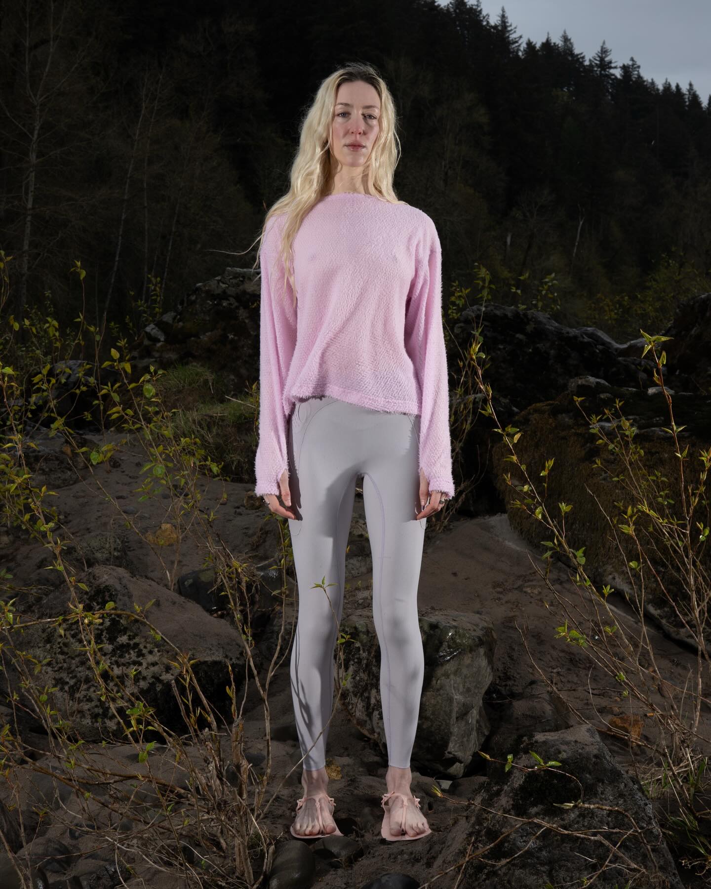 A blonde woman wearing a pink sweater and grey leggings stands among rocky terrain and sparse shrubs against a backdrop of dark evergreen trees.
