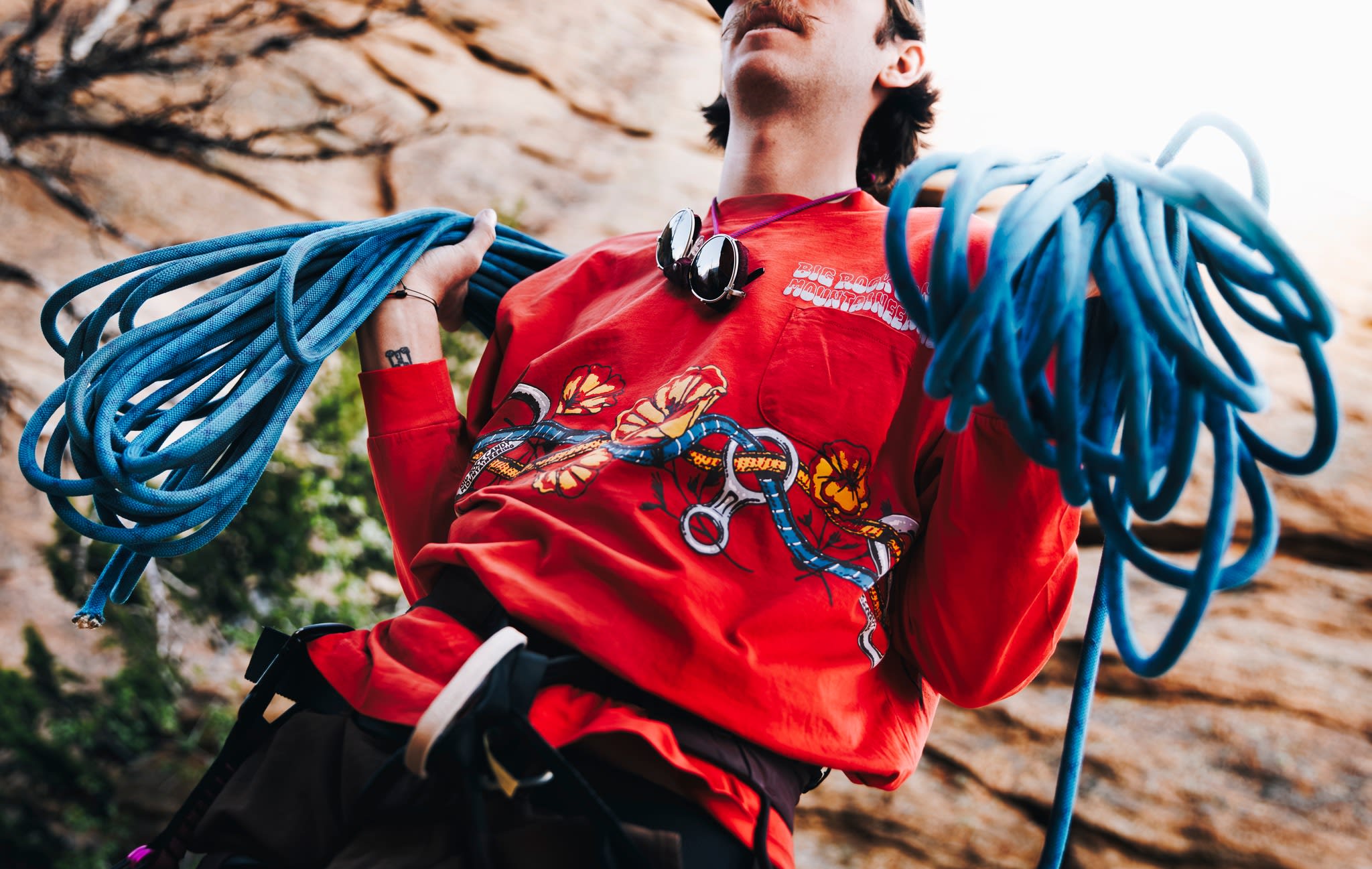 A man in a red sweatshirt holds coiled blue ropes in the foreground, with a rocky cliff face in the background.