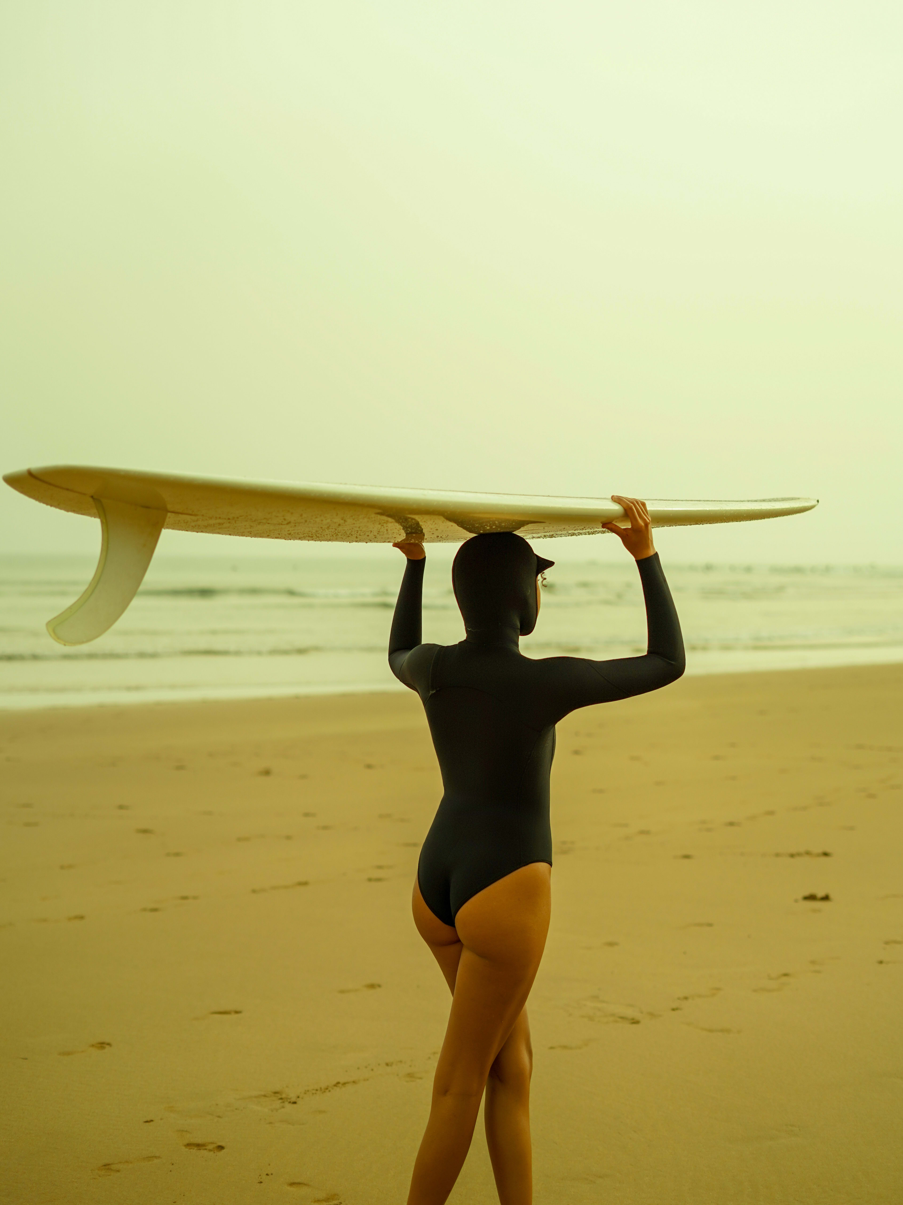 A person in a black bodysuit holds a surfboard over their head on a sandy beach with ocean waves in the background.