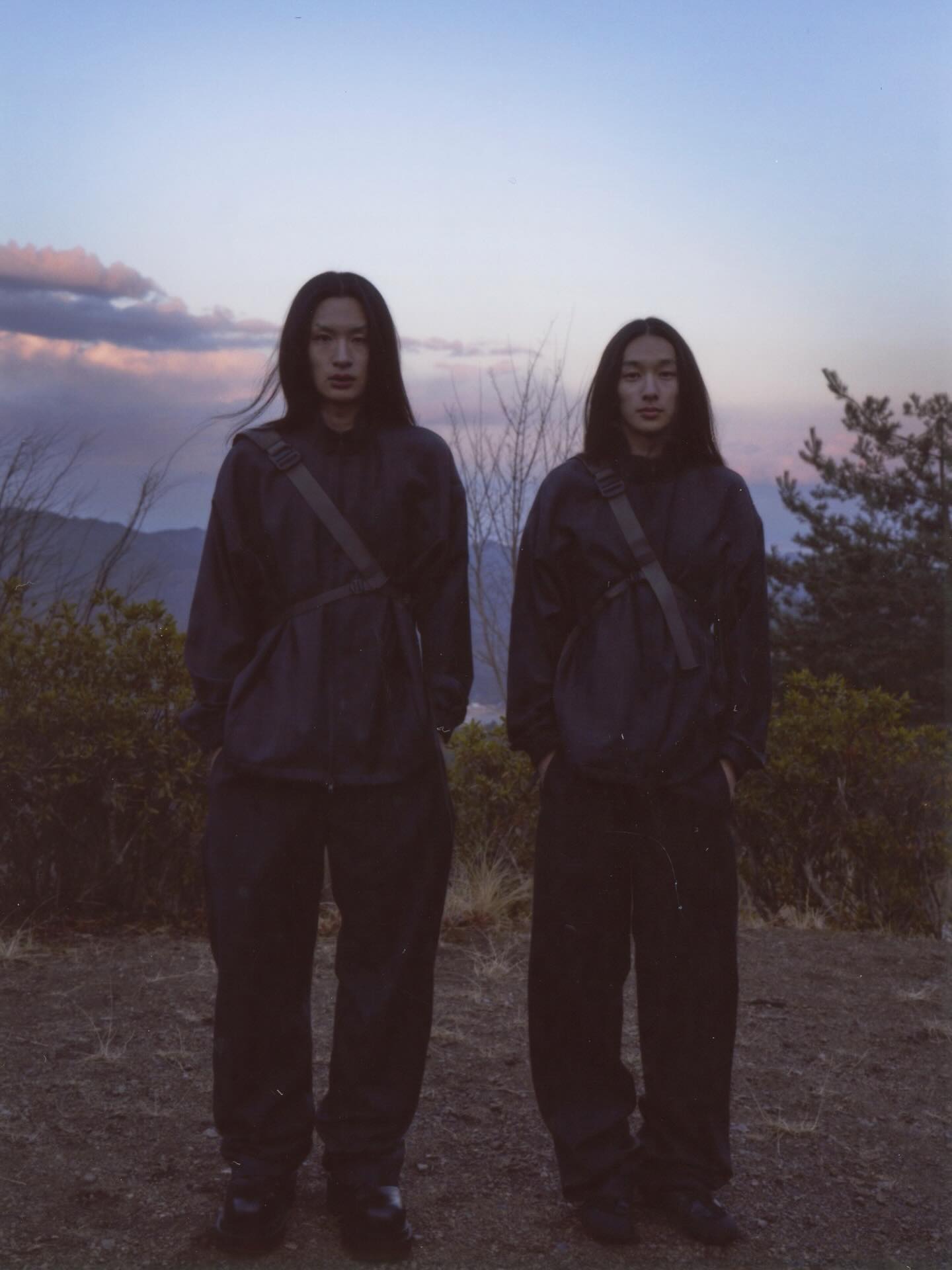 Two people dressed in dark, oversized outfits stand in a grassy foreground against a backdrop of rolling hills and a twilight sky.