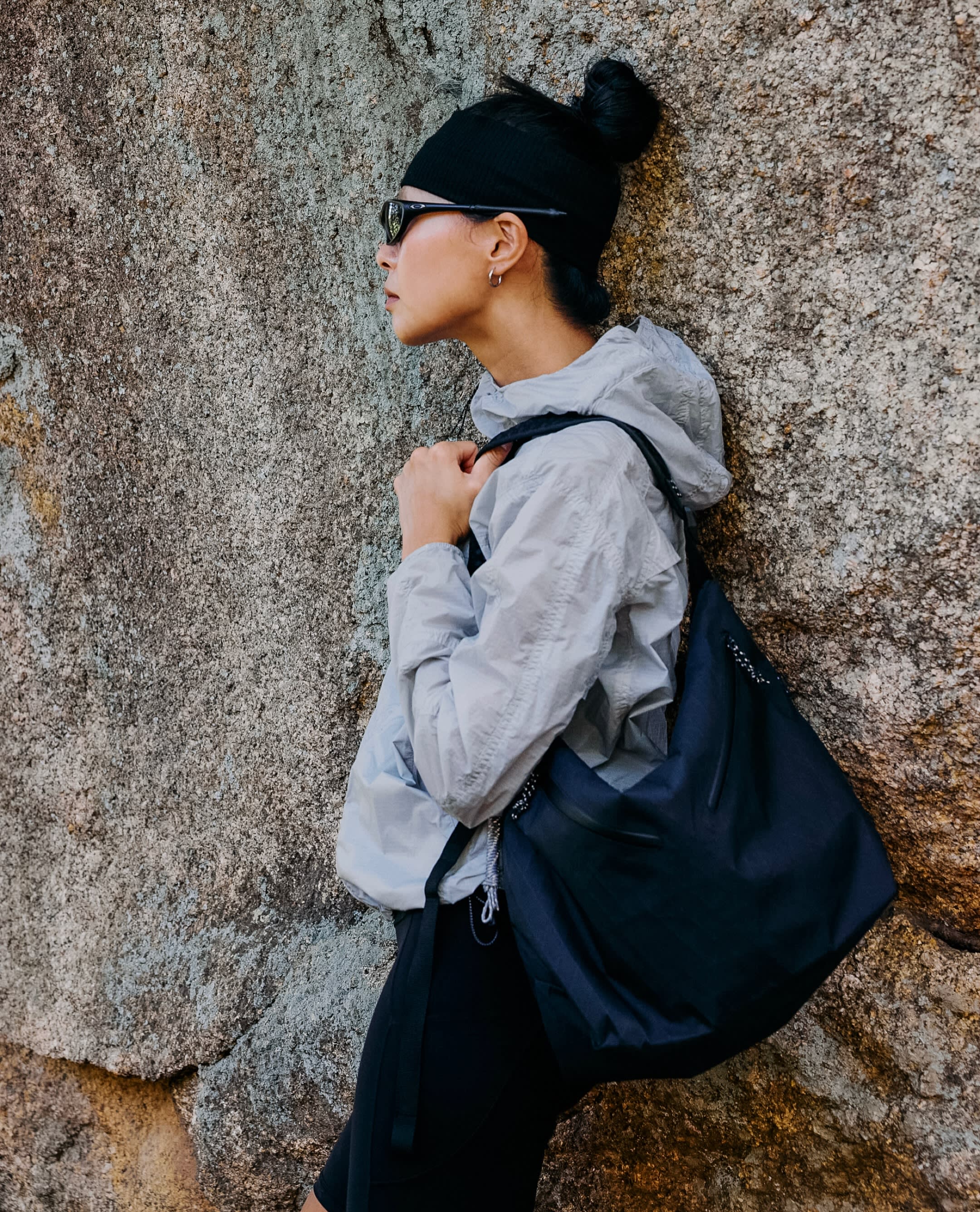 A woman wearing a black beanie, sunglasses, and a light grey jacket leans against a textured grey rock wall while carrying a large black bag.