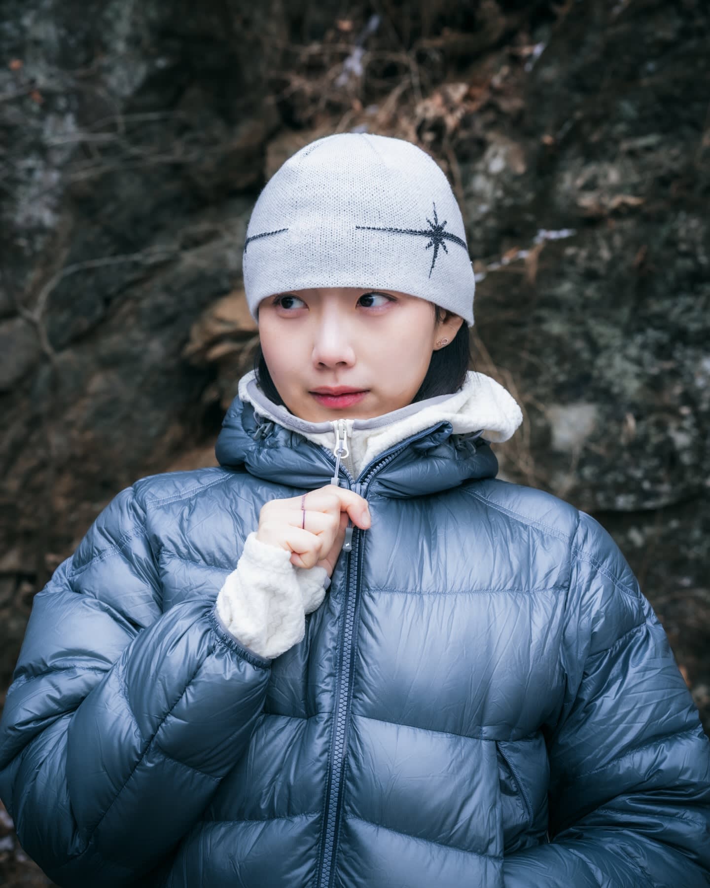 A woman wearing a grey beanie and a blue puffer jacket stands against a dark, rocky background.