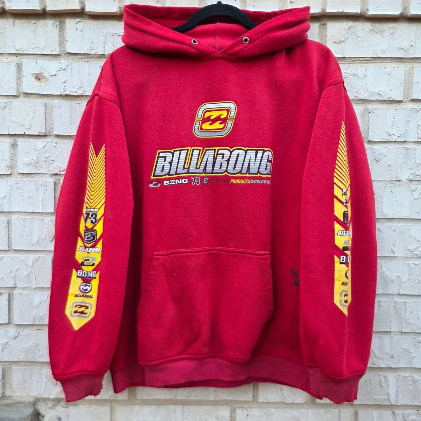 A red Billabong hoodie with yellow graphics hangs against a white brick wall.