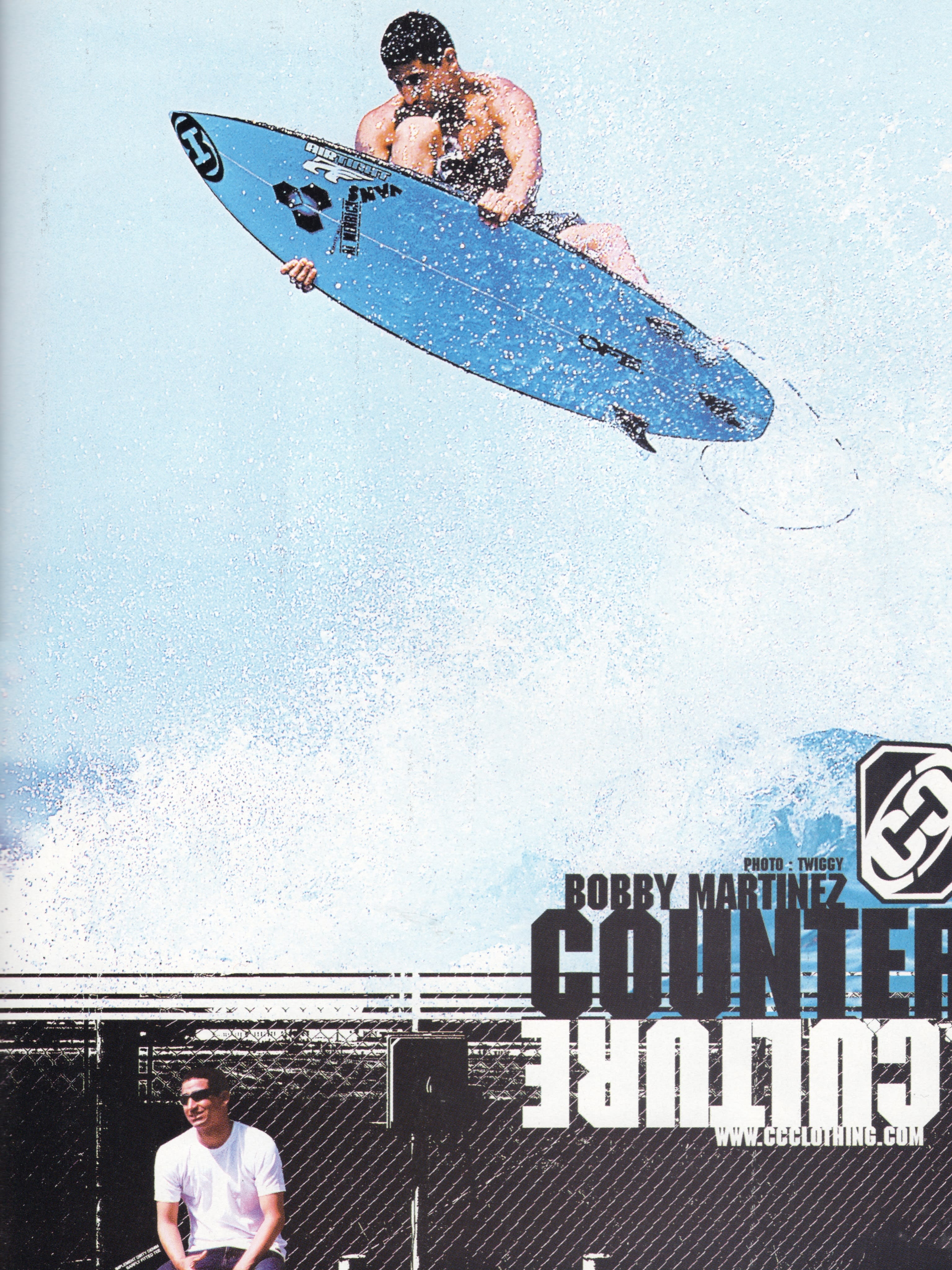 A man holds a blue surfboard amidst splashing water in the upper foreground, while another man stands before a black fence and bold text in the lower background.