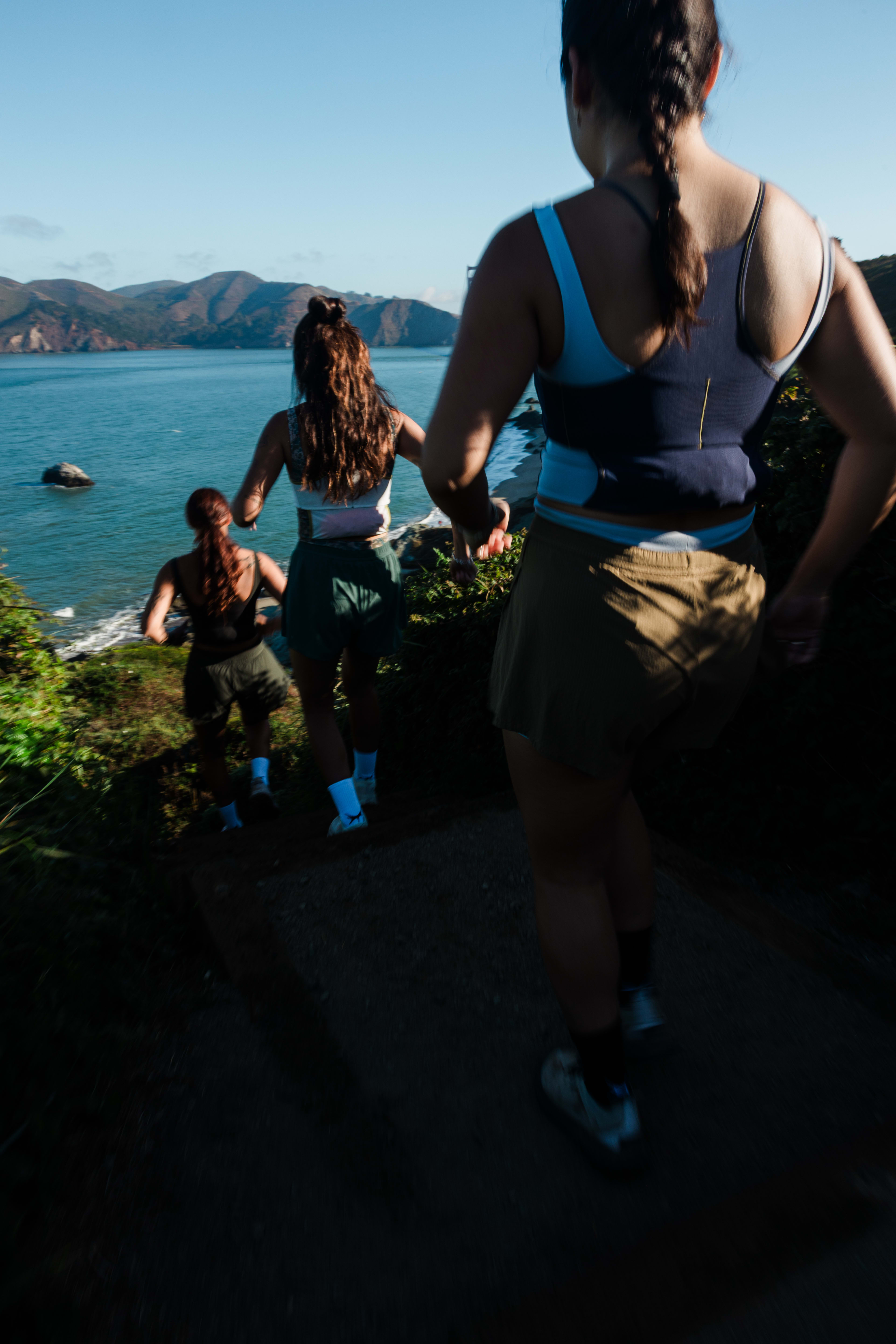 Three women in athletic wear run along a coastal path toward a scenic view of the ocean and distant mountains.
