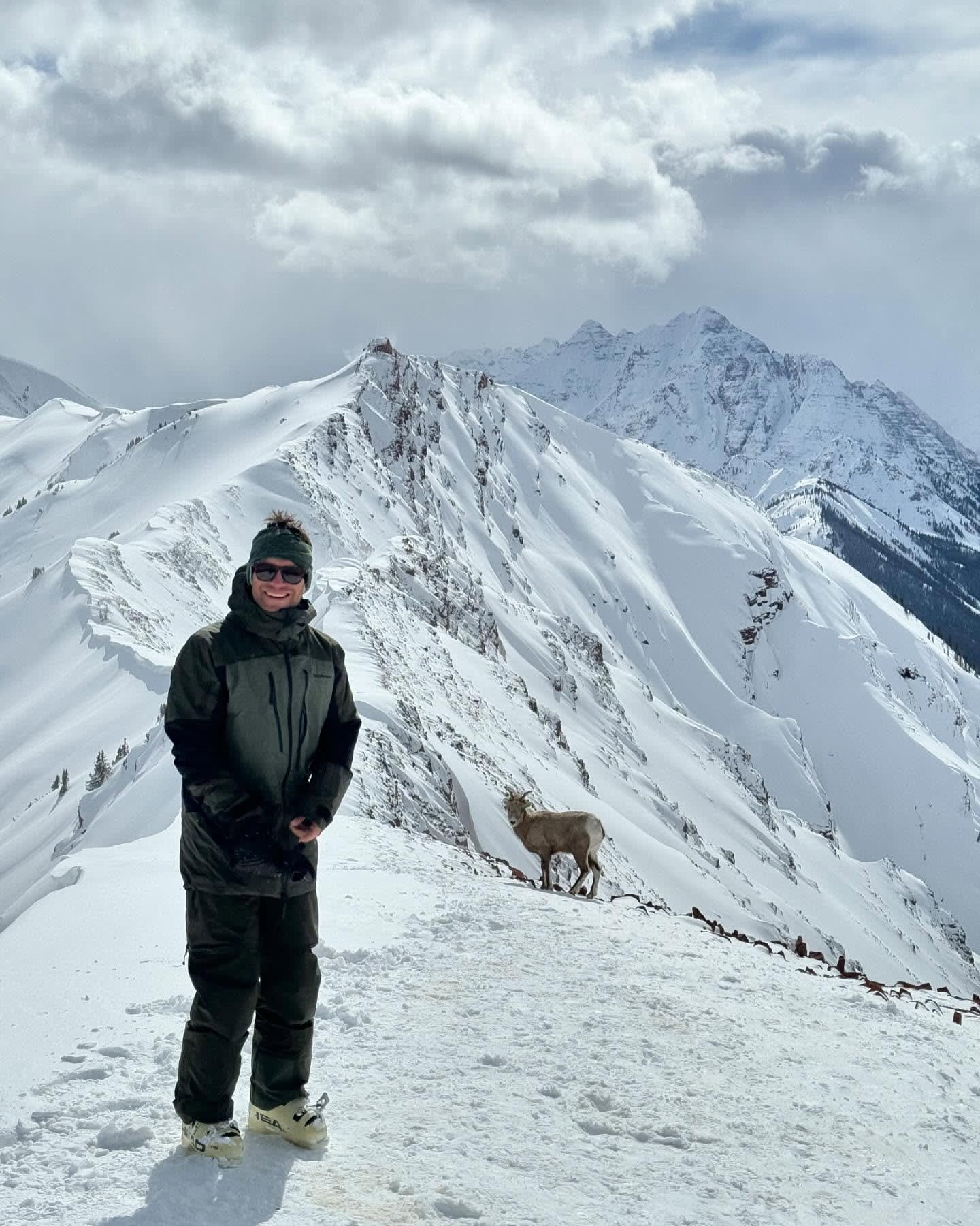 A man in dark winter gear stands on a snowy ridge with a mountain goat nearby, set against a backdrop of jagged, snow-covered peaks and a cloudy sky.