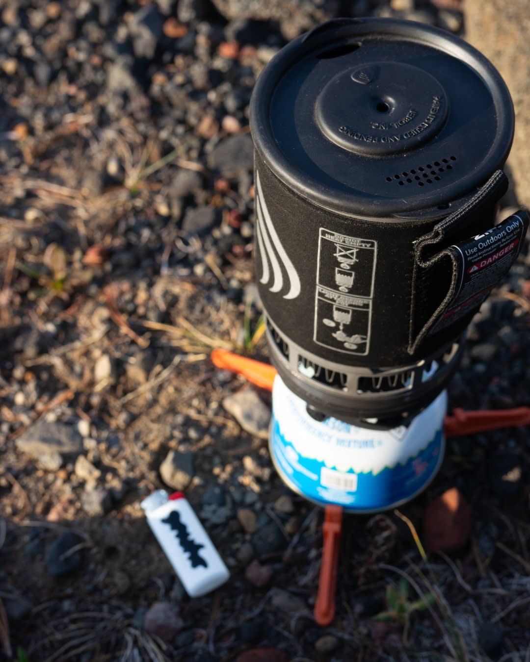 A portable camping stove and lighter sit on a rocky, earthy ground.