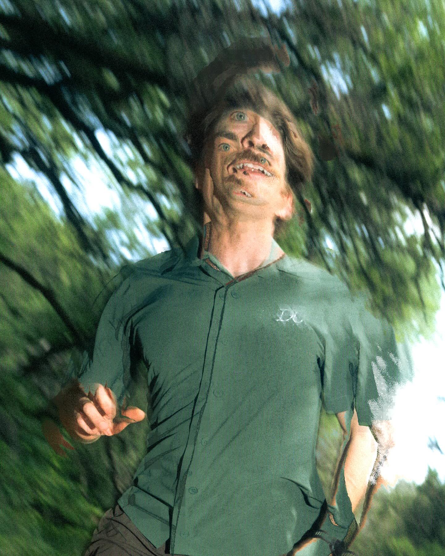 A man with a distorted face wearing a green shirt stands against a blurred background of green trees.