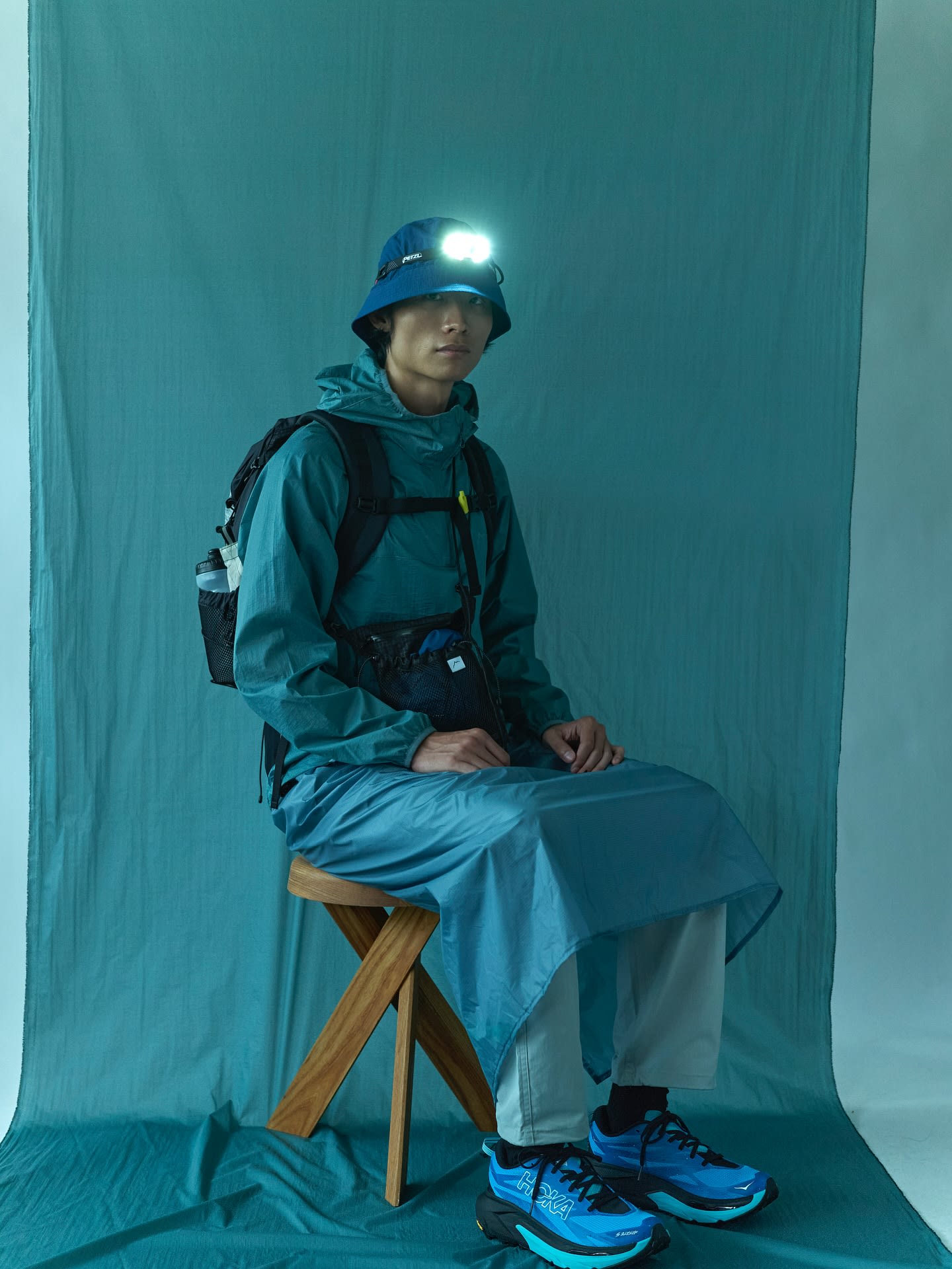 A man wearing a blue outdoor outfit and a lit headlamp sits on a wooden stool against a draped teal fabric backdrop.