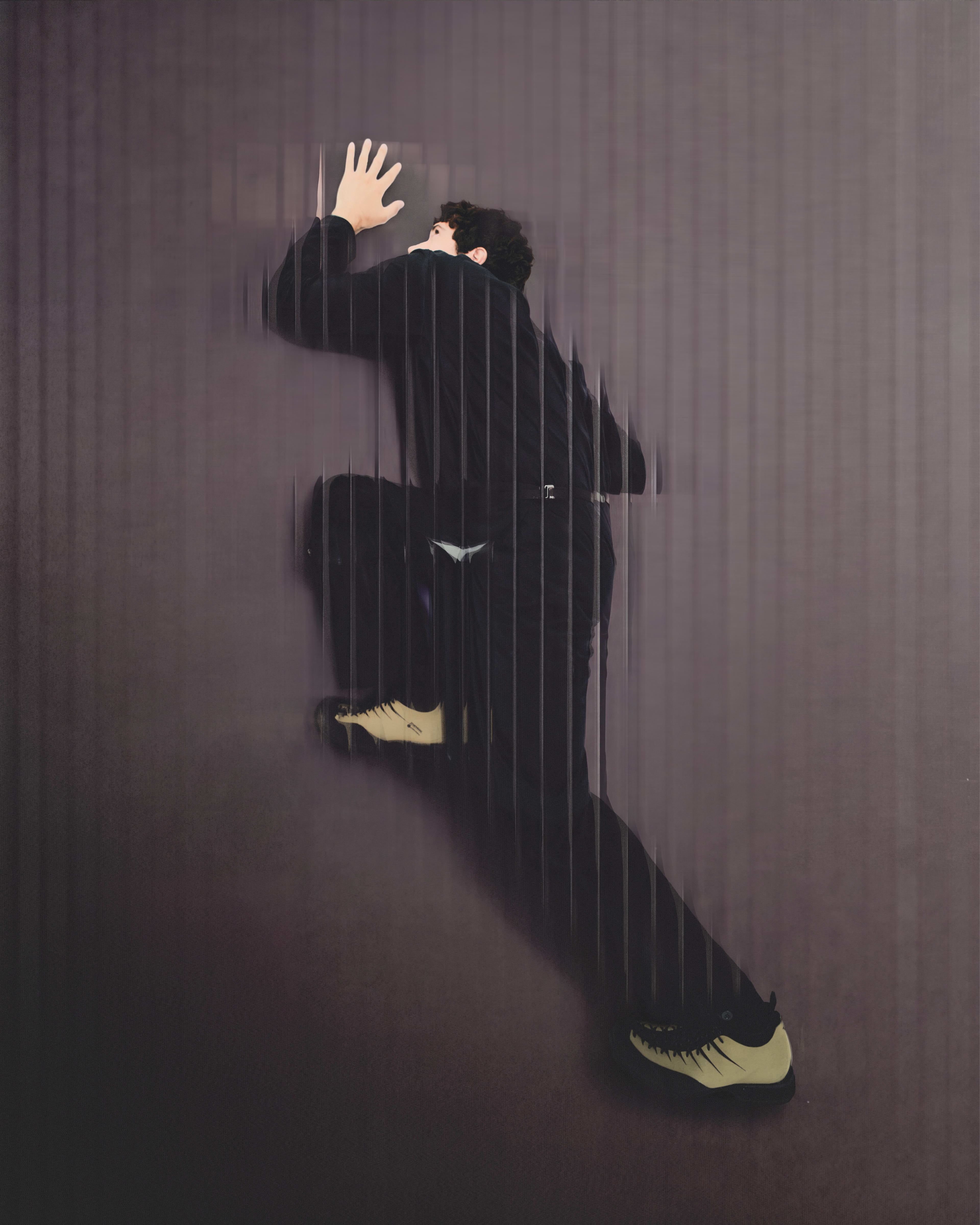 A man in a black suit and yellow sneakers presses his hand against a dark, vertically ribbed wall.