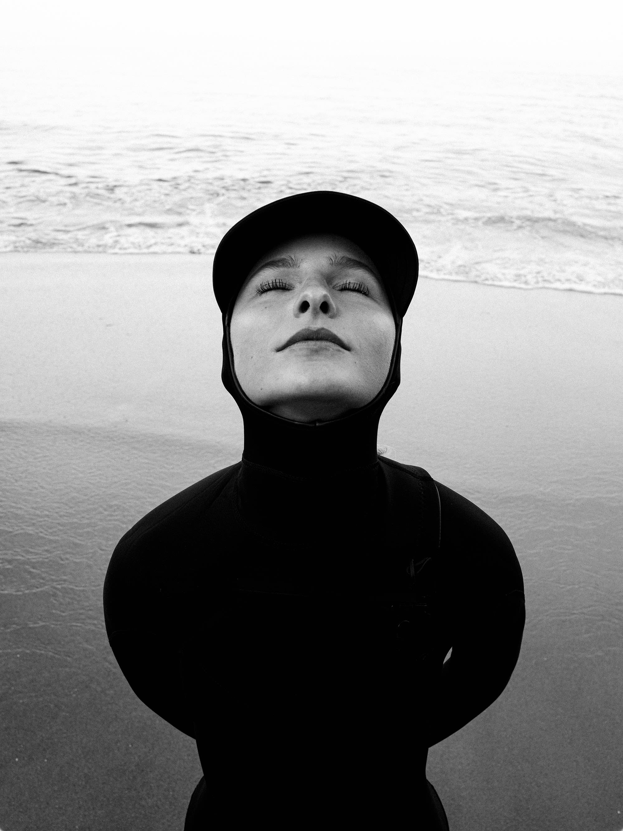 A person in a black wetsuit and hood stands with eyes closed on a sandy beach with ocean waves in the background.