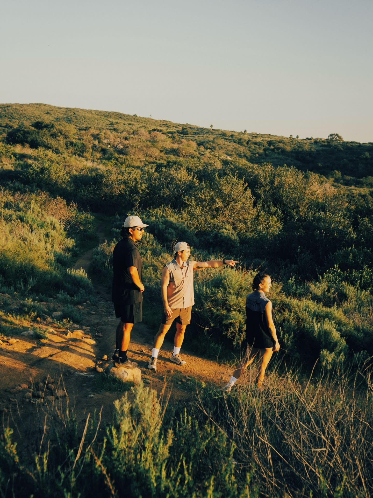 Three people stand on a dirt path in the foreground, with one person pointing toward a vast, rolling green hillside in the background.