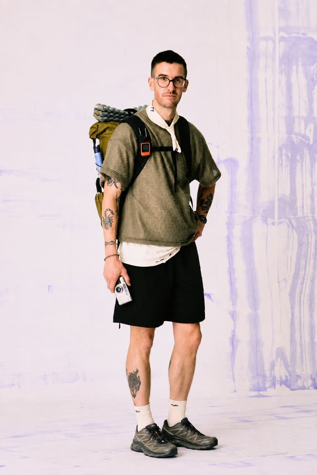 A man wearing glasses, a textured olive shirt, black shorts, and a large hiking backpack stands against a white background with purple paint streaks.