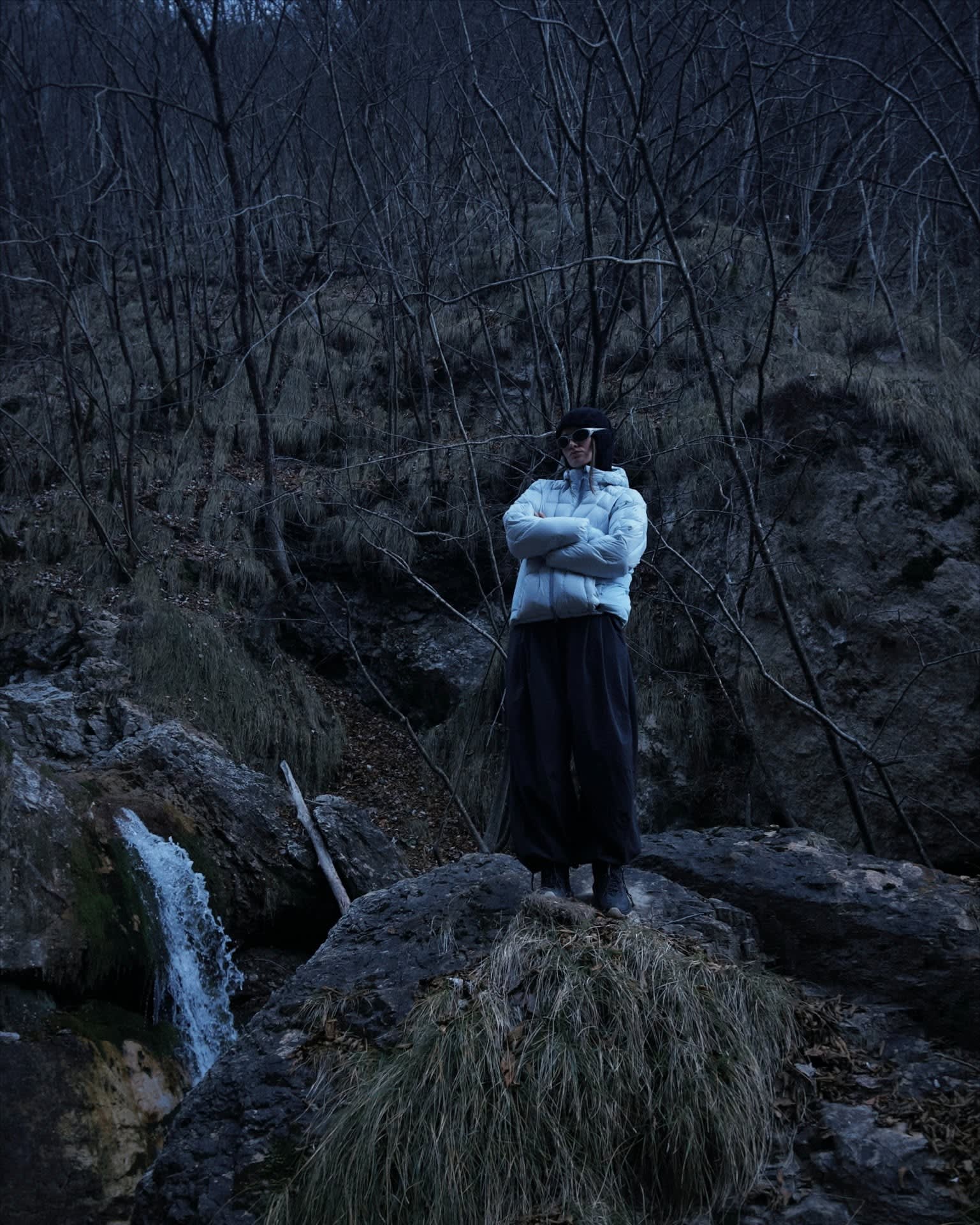 A person wearing a white puffer jacket and wide black trousers stands on a rocky ledge in the foreground, set against a background of dark, leafless trees and a small waterfall.