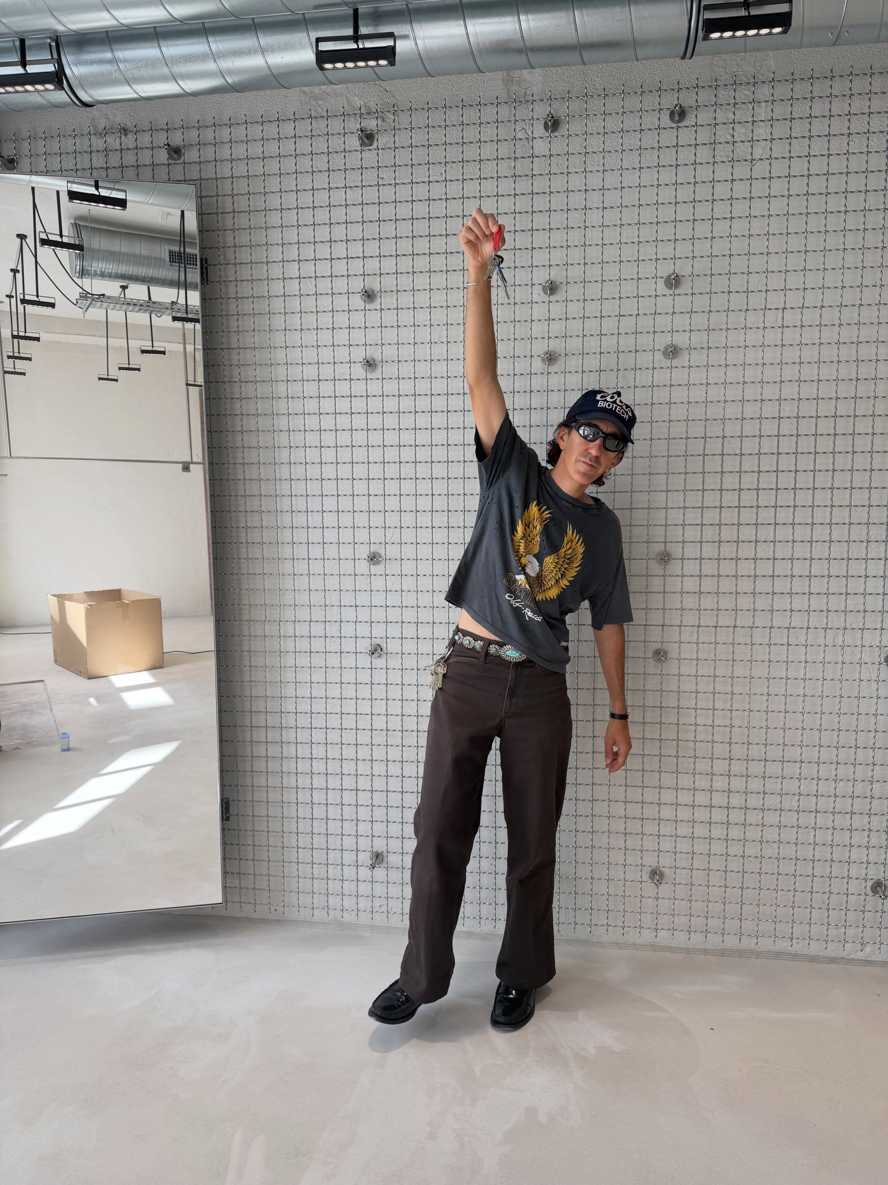 A person wearing a graphic t-shirt, brown pants, and sunglasses stands with one arm raised against a grid-patterned wall in a minimalist room.
