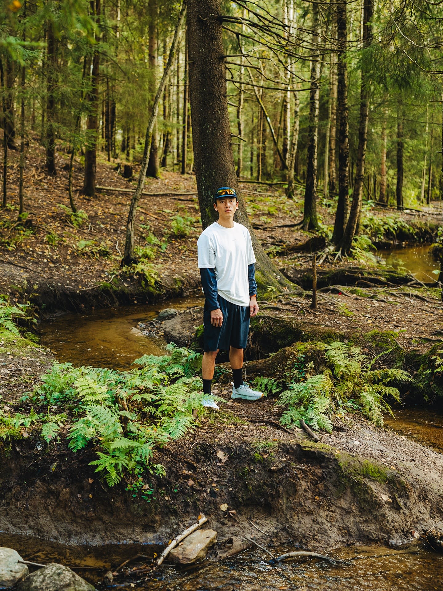 A man in a white t-shirt and black shorts stands on a mossy riverbank surrounded by a dense forest of tall trees.
