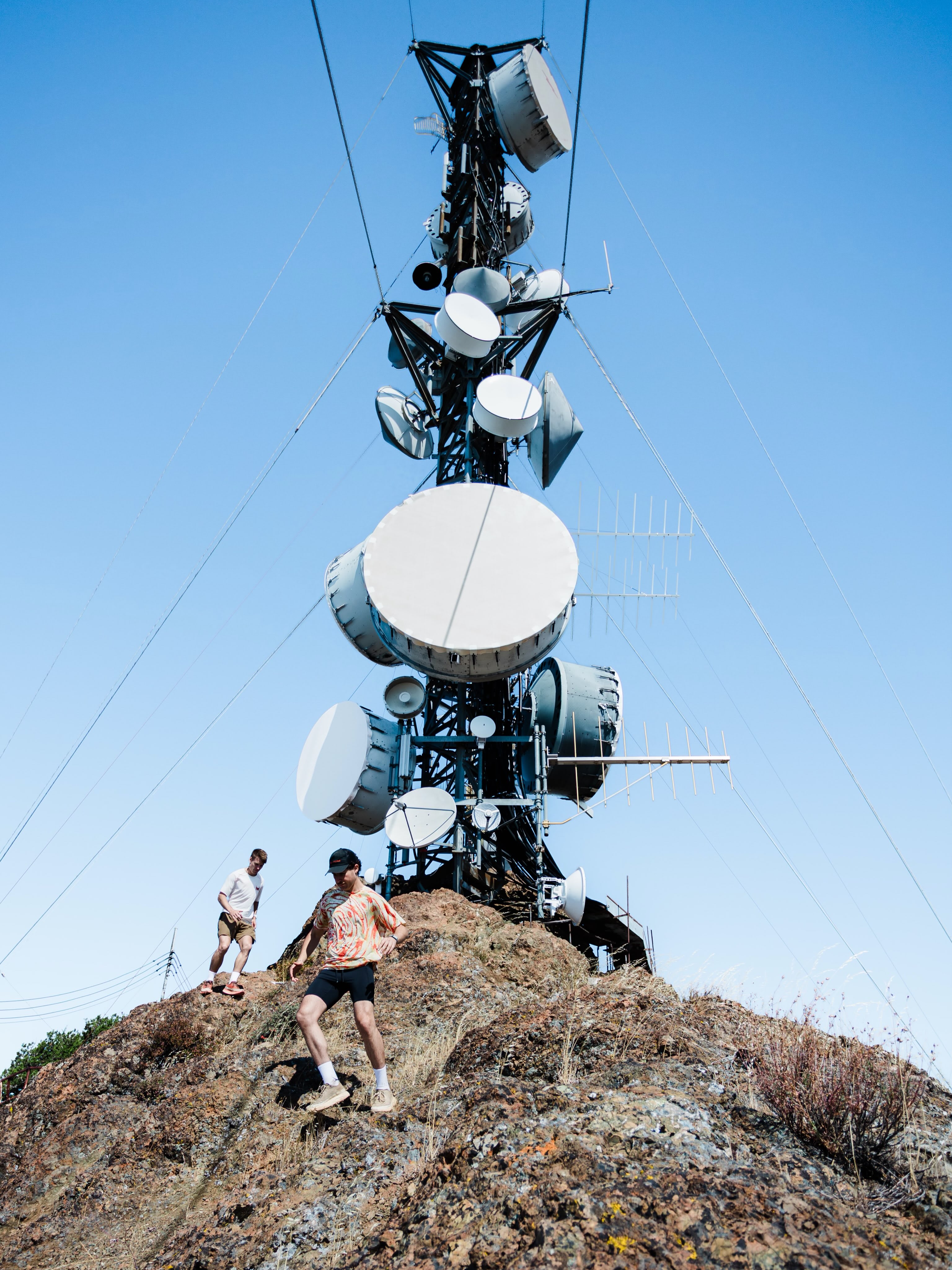 Two men hike up a rocky hillside in the foreground, with a tall telecommunications tower standing against a clear blue sky in the background.