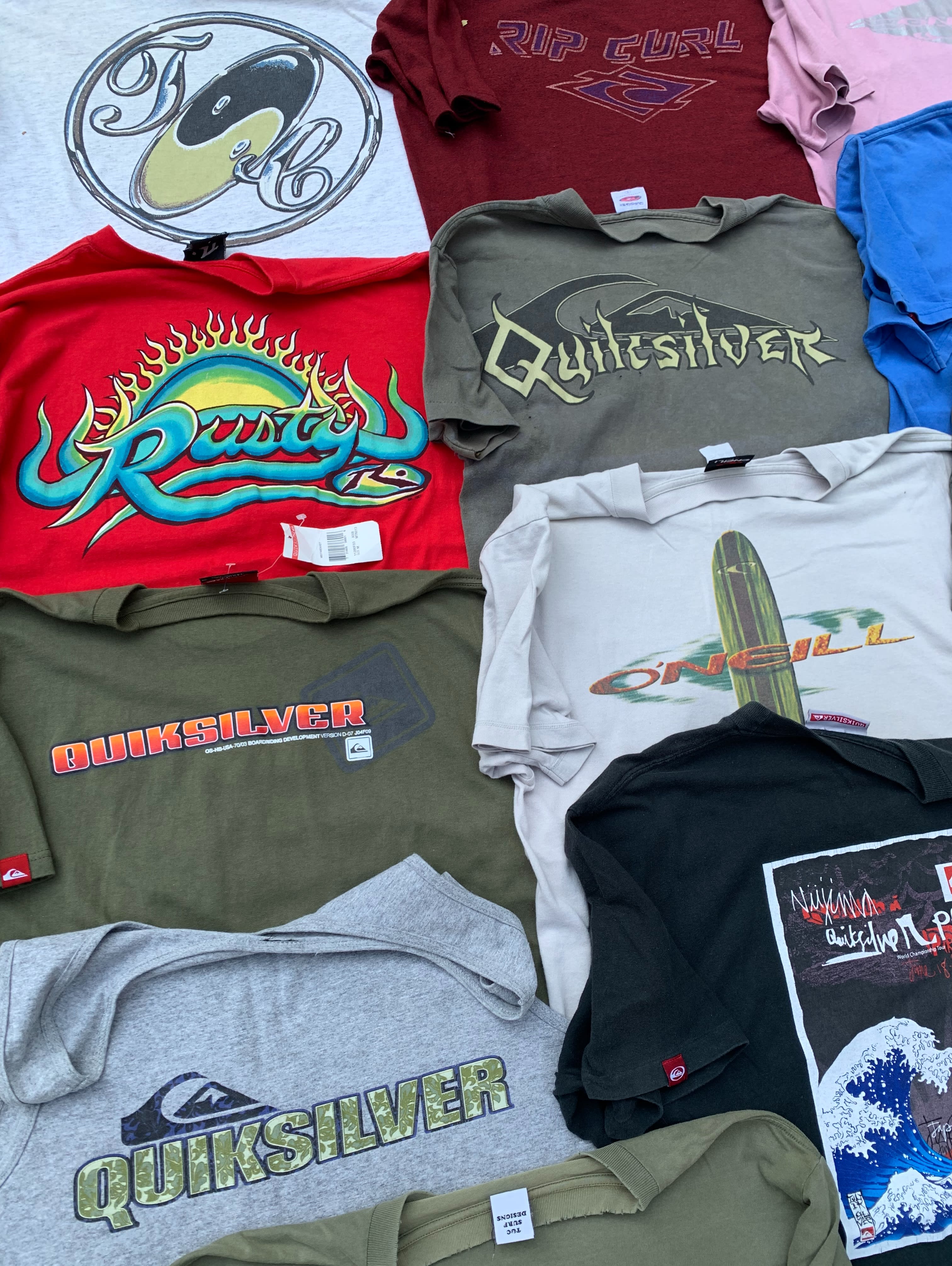 A collection of colorful surf-brand t-shirts is spread across a plain white background.
