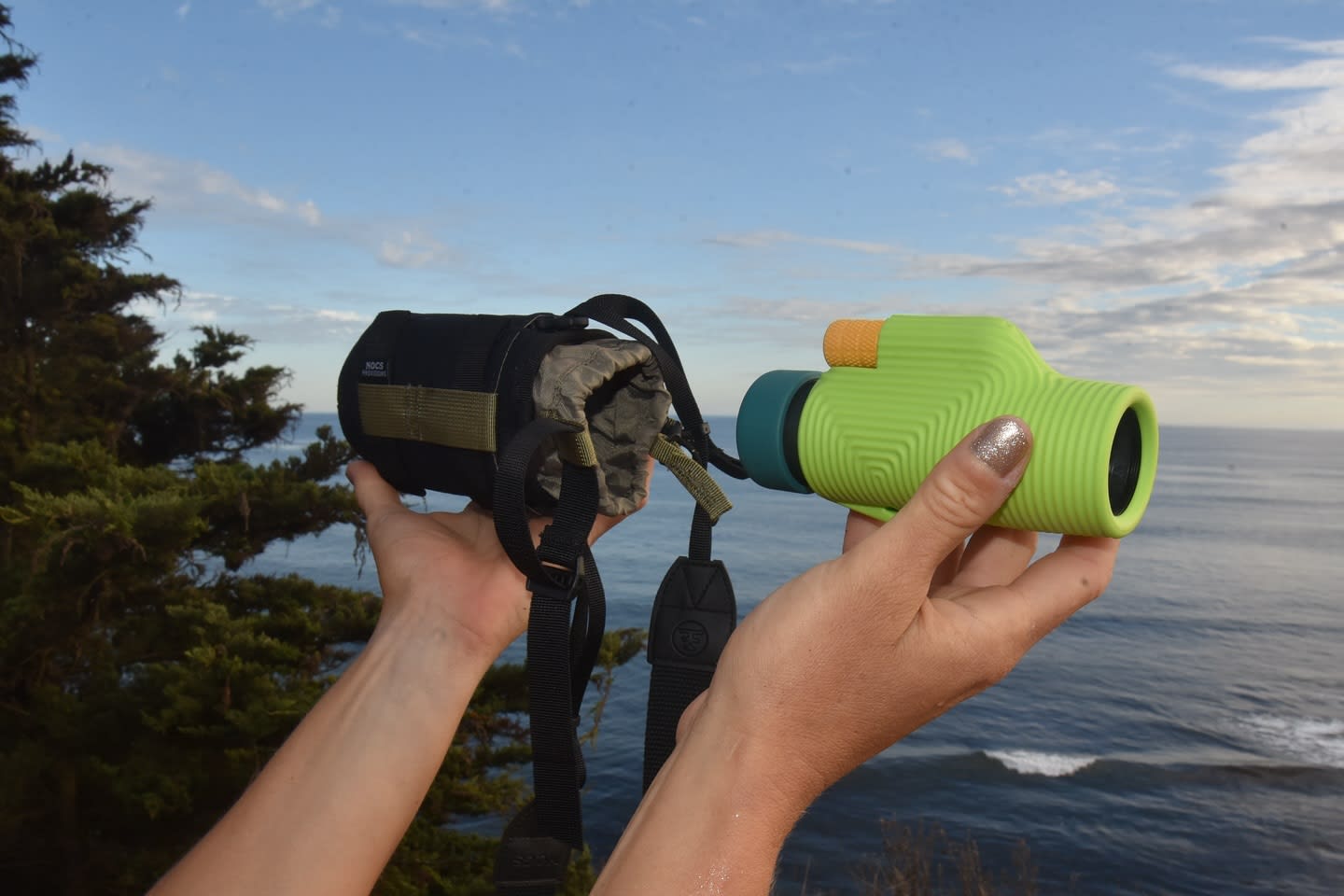 Hands hold a green toy binoculars and its black case against a backdrop of the ocean and sky.