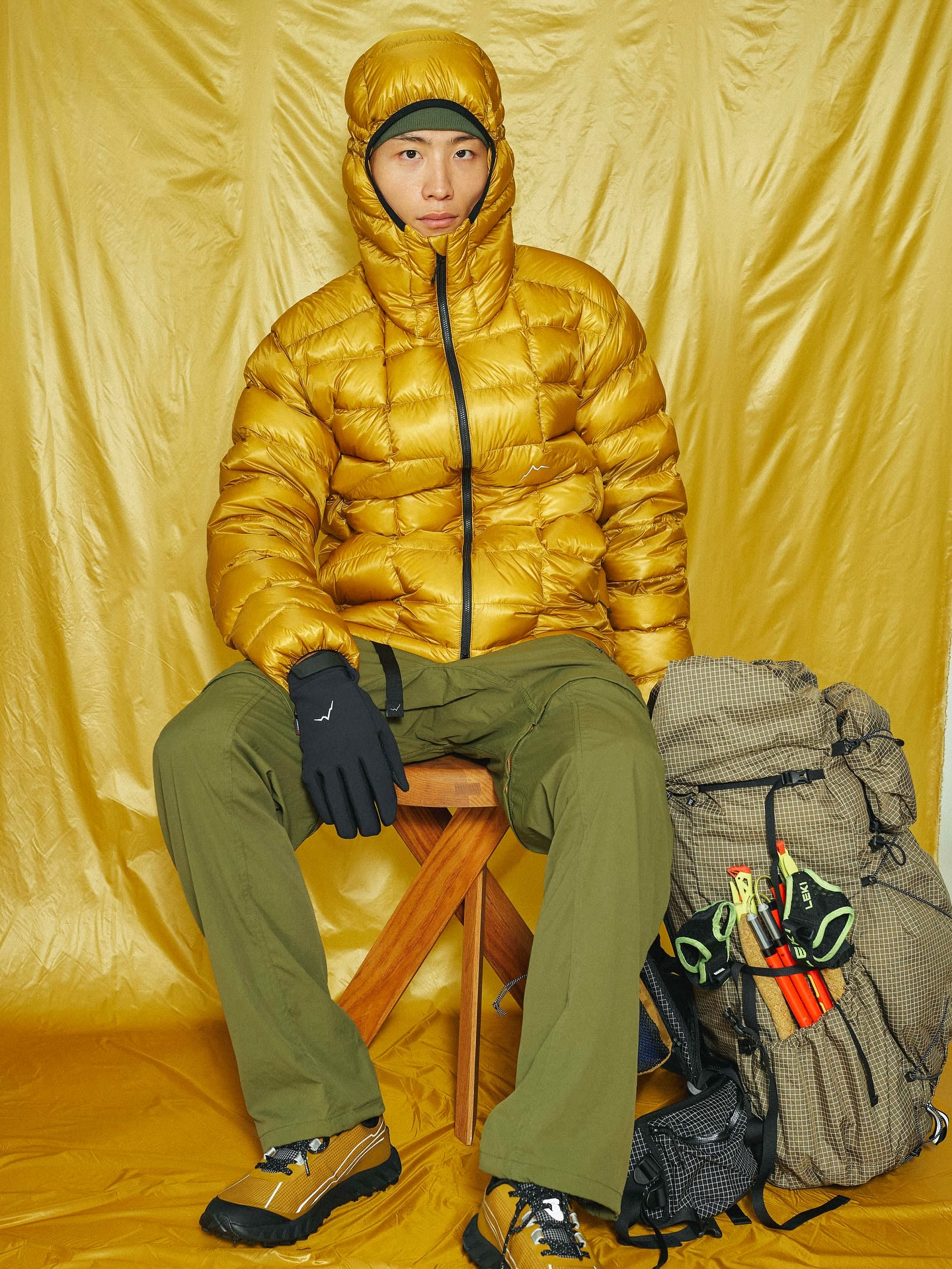 A man wearing a gold puffer jacket and olive green pants sits on a wooden stool next to a hiking backpack against a shimmering gold fabric backdrop.
