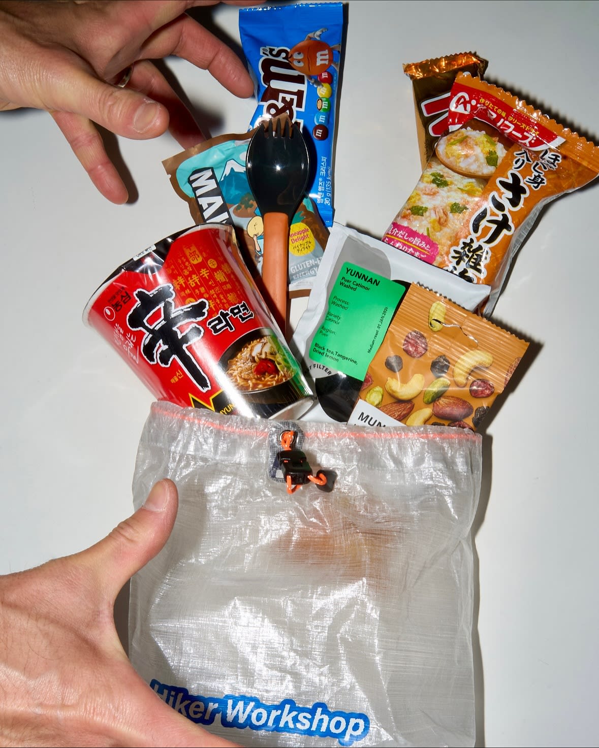 Various packaged snacks and a cup of noodles are scattered around a clear plastic bag on a white surface.
