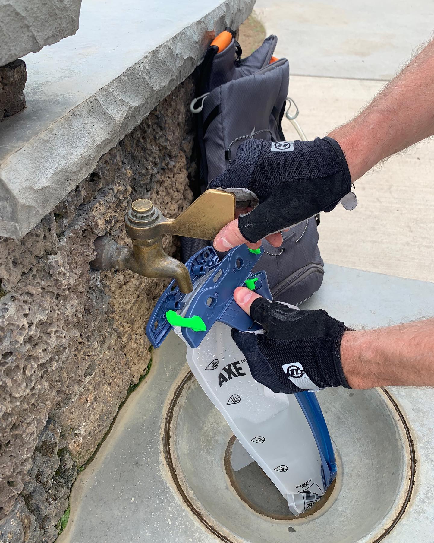 A person wearing black gloves fills a blue and white AXE water bottle from a brass outdoor faucet against a stone wall and grey bag.