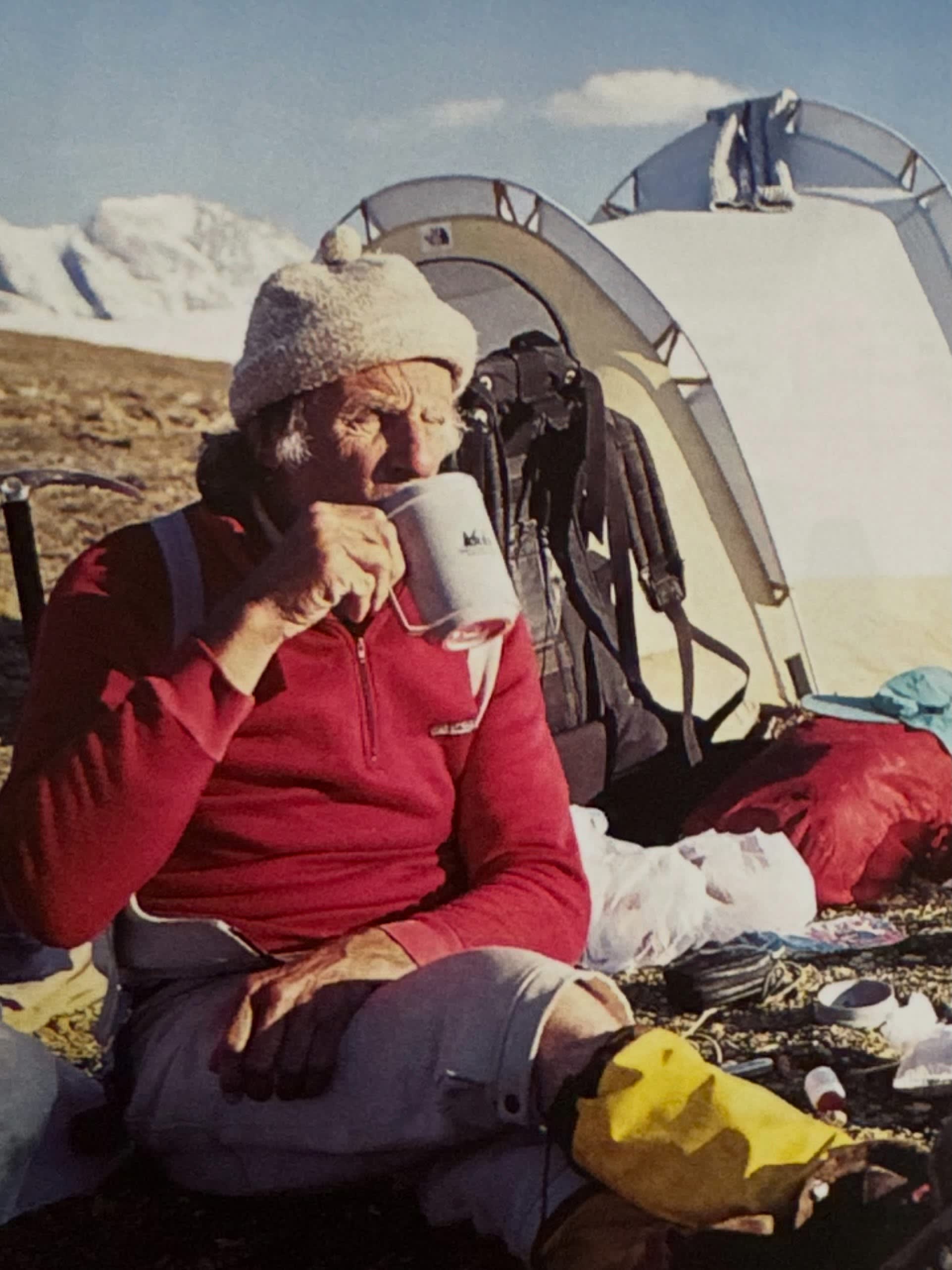A man in a red shirt and white hat drinks from a mug in the foreground, while a tent and snow-capped mountains are visible in the background.