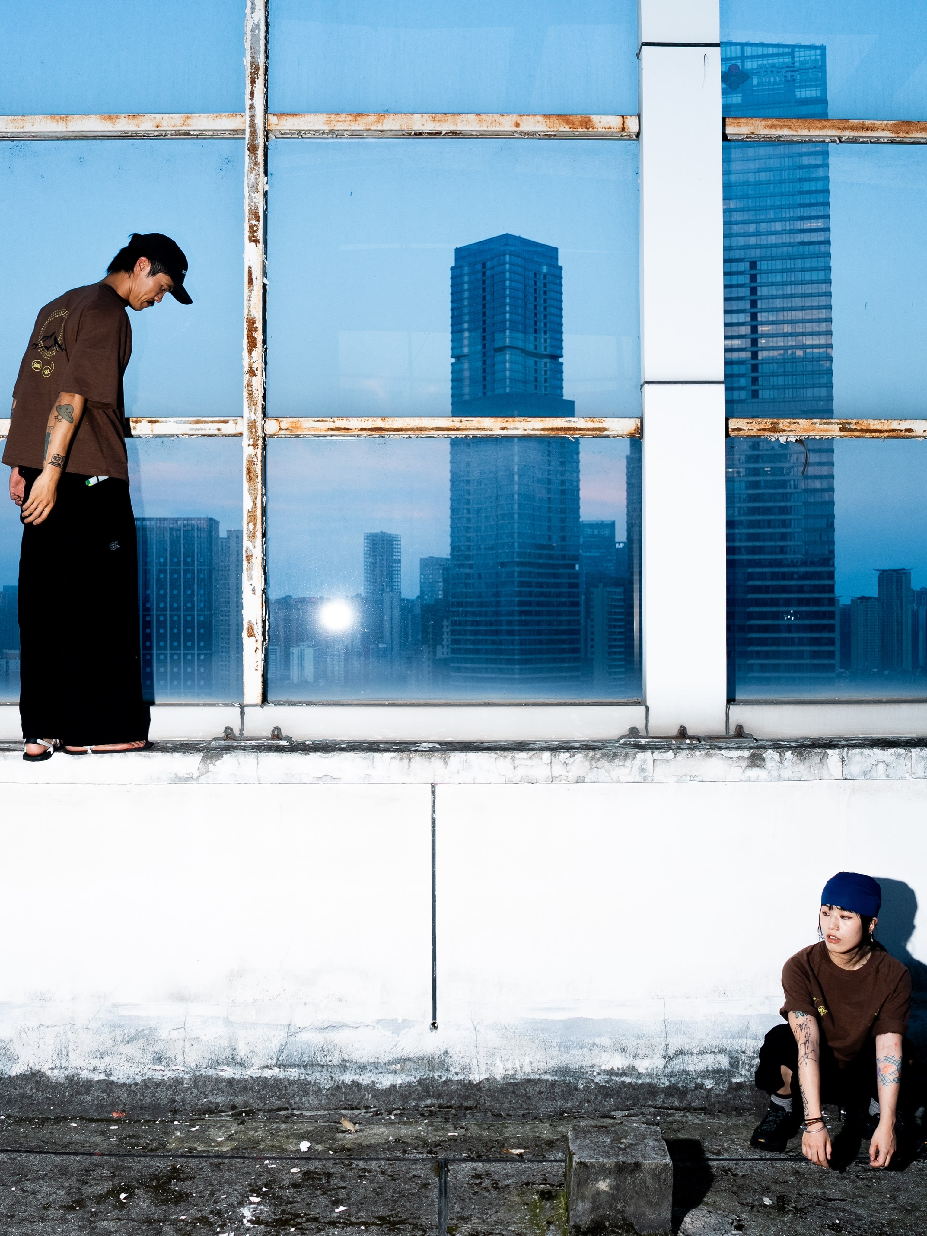 Two people in brown shirts and hats are positioned against a white wall and large blue glass windows reflecting a city skyline.