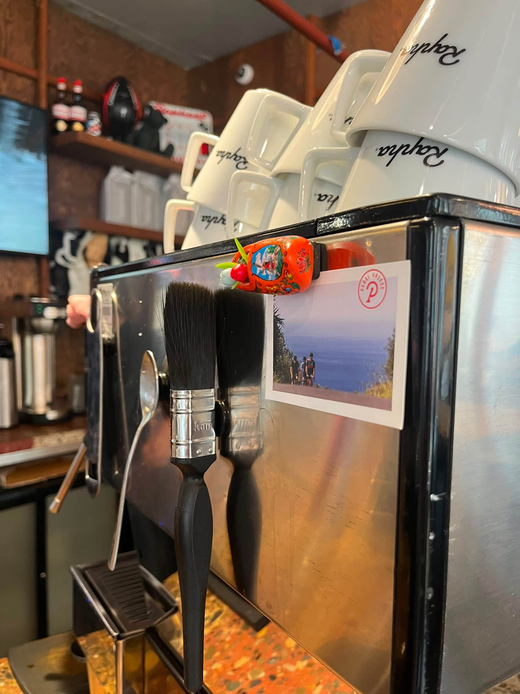 Two paintbrushes hang in front of a stainless steel espresso machine topped with stacked coffee cups, while shelves with various items are visible in the background.