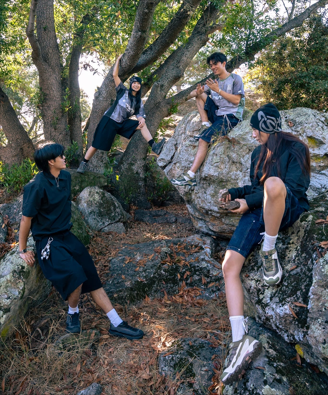 Four young people in casual attire relax among rocky outcrops and trees in a wooded area.