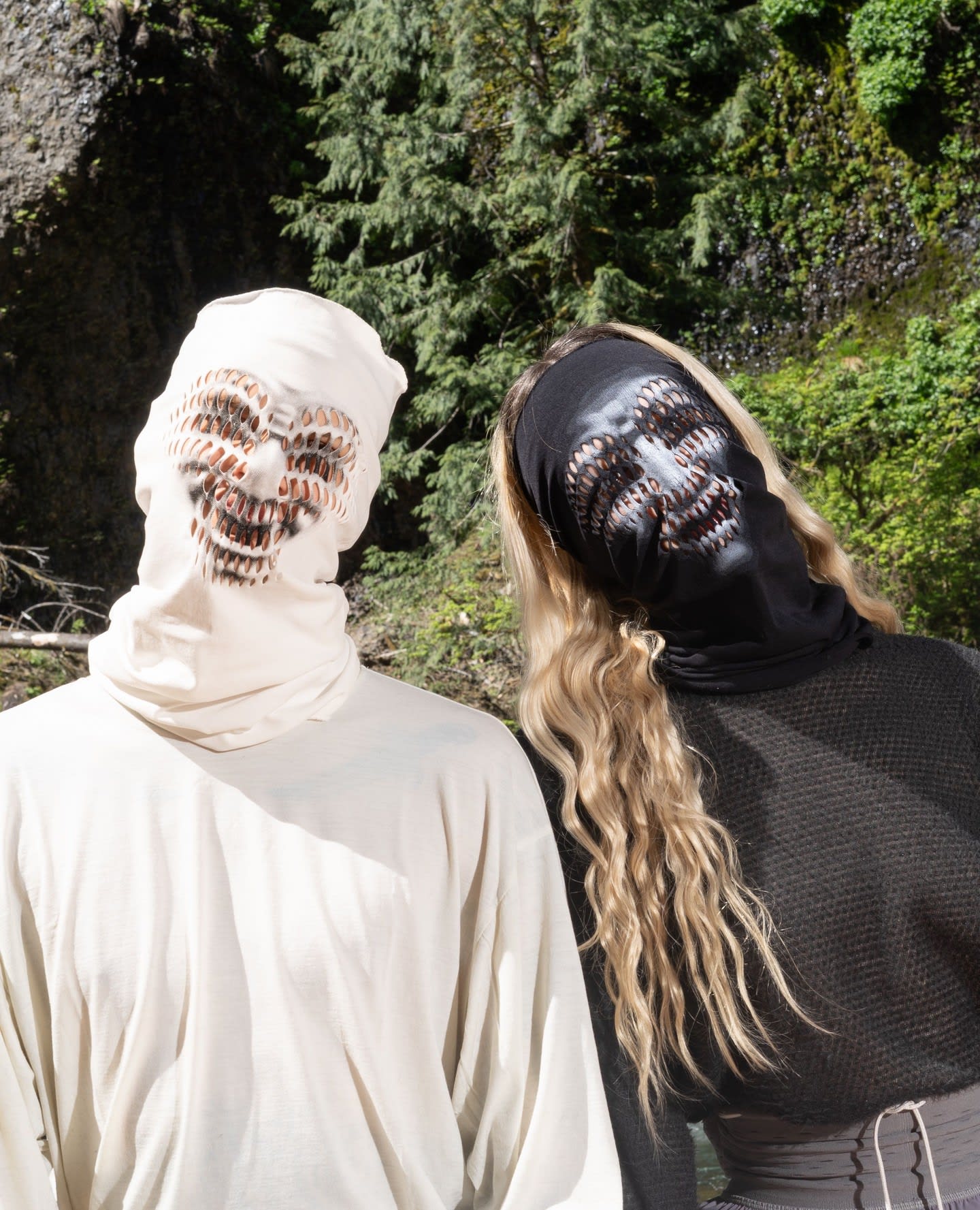 Two people wearing skull-patterned face masks in white and black stand before a backdrop of lush green trees and rocky terrain.