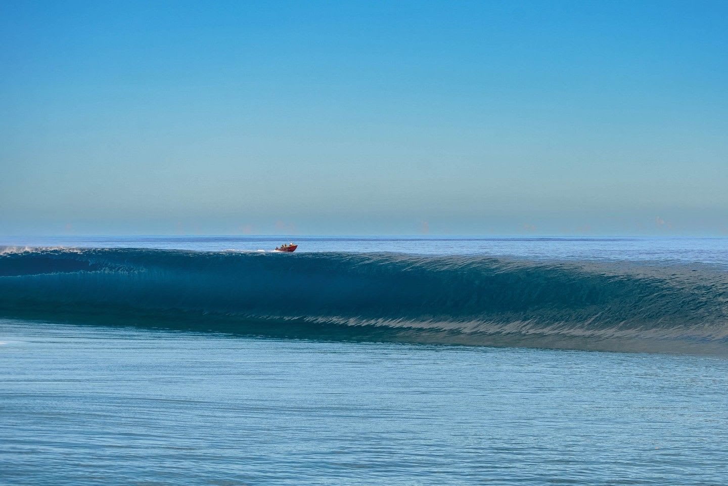 A large, smooth ocean wave forms with a small red boat visible on the horizon.