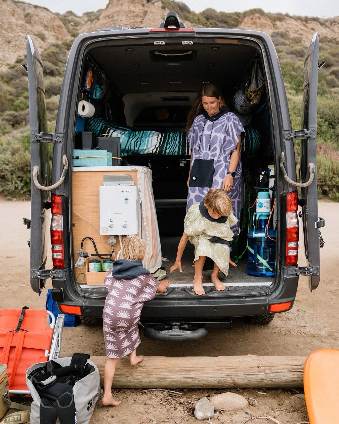 A woman and two boys in patterned surf ponchos get into the open back of a camper van on a dirt road, with hills in the distance.