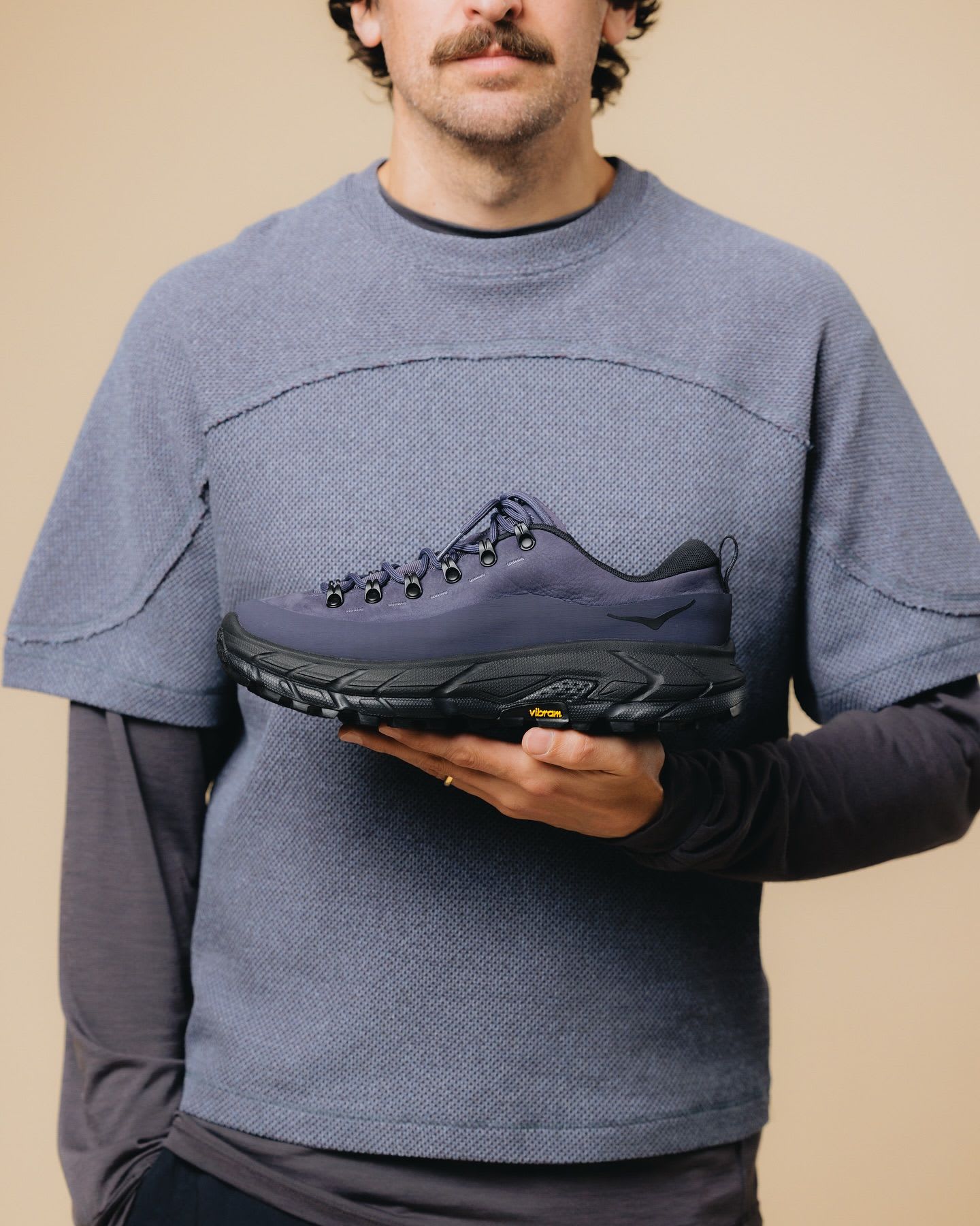 Man with a mustache holding a dark blue Hoka hiking shoe with a black Vibram sole.