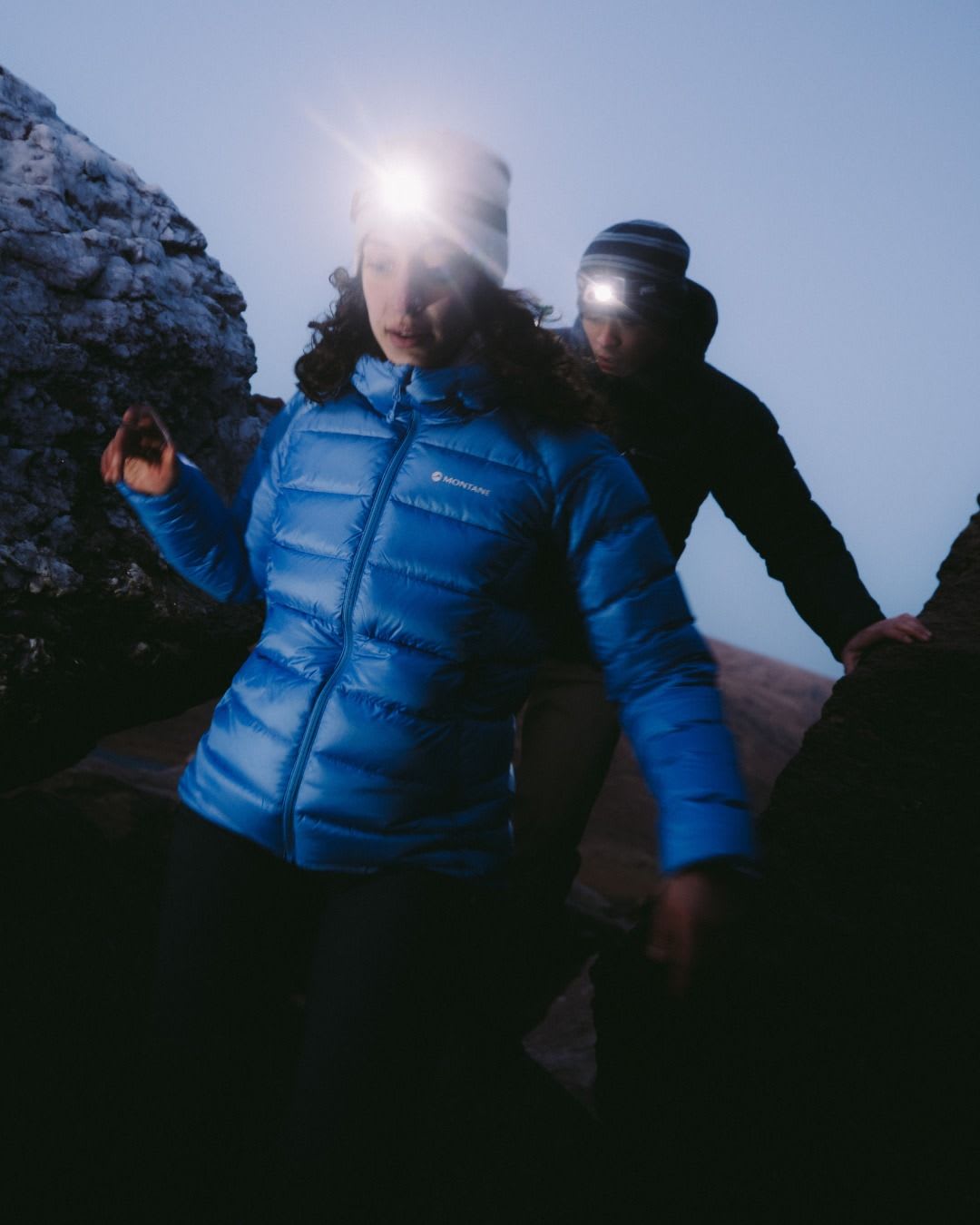 Two hikers wearing bright headlamps navigate dark, rocky terrain.