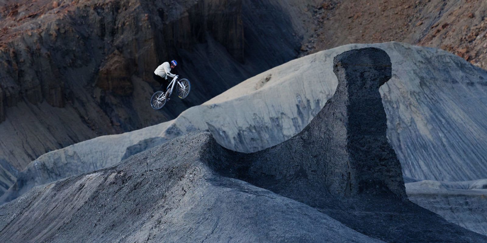 A mountain biker jumps over a dirt hill in a rocky, barren landscape.